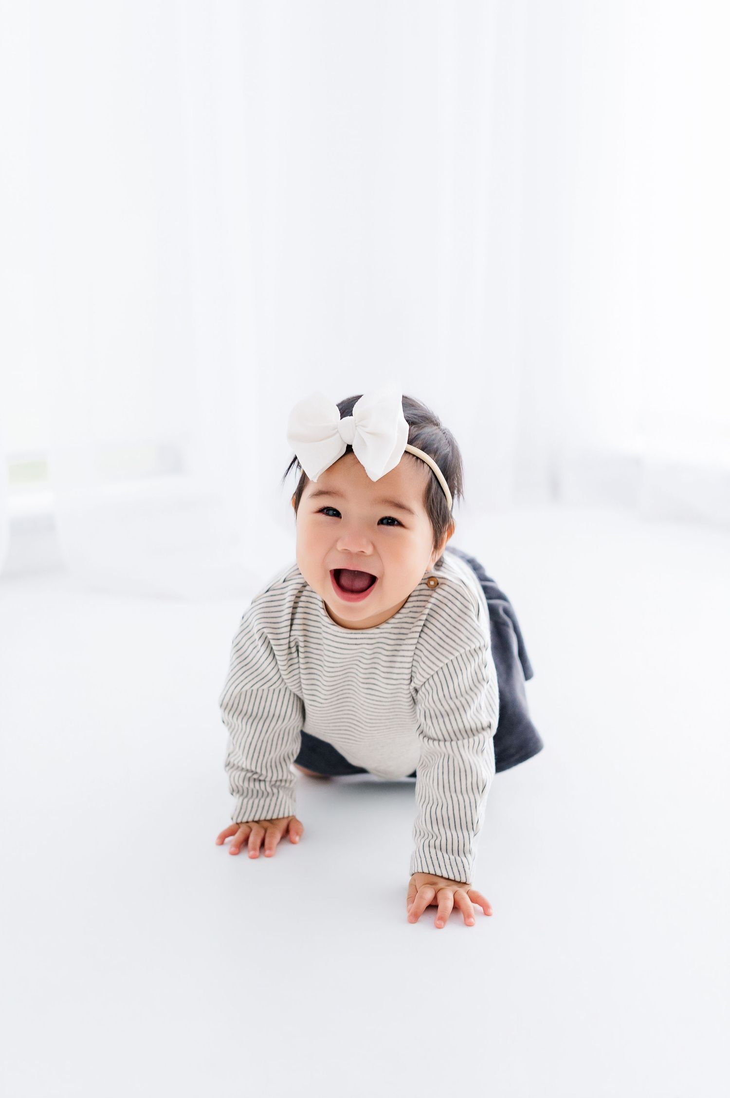A happy baby girl in a striped sweater and bow headband crawling on a white floor, smiling joyfully.