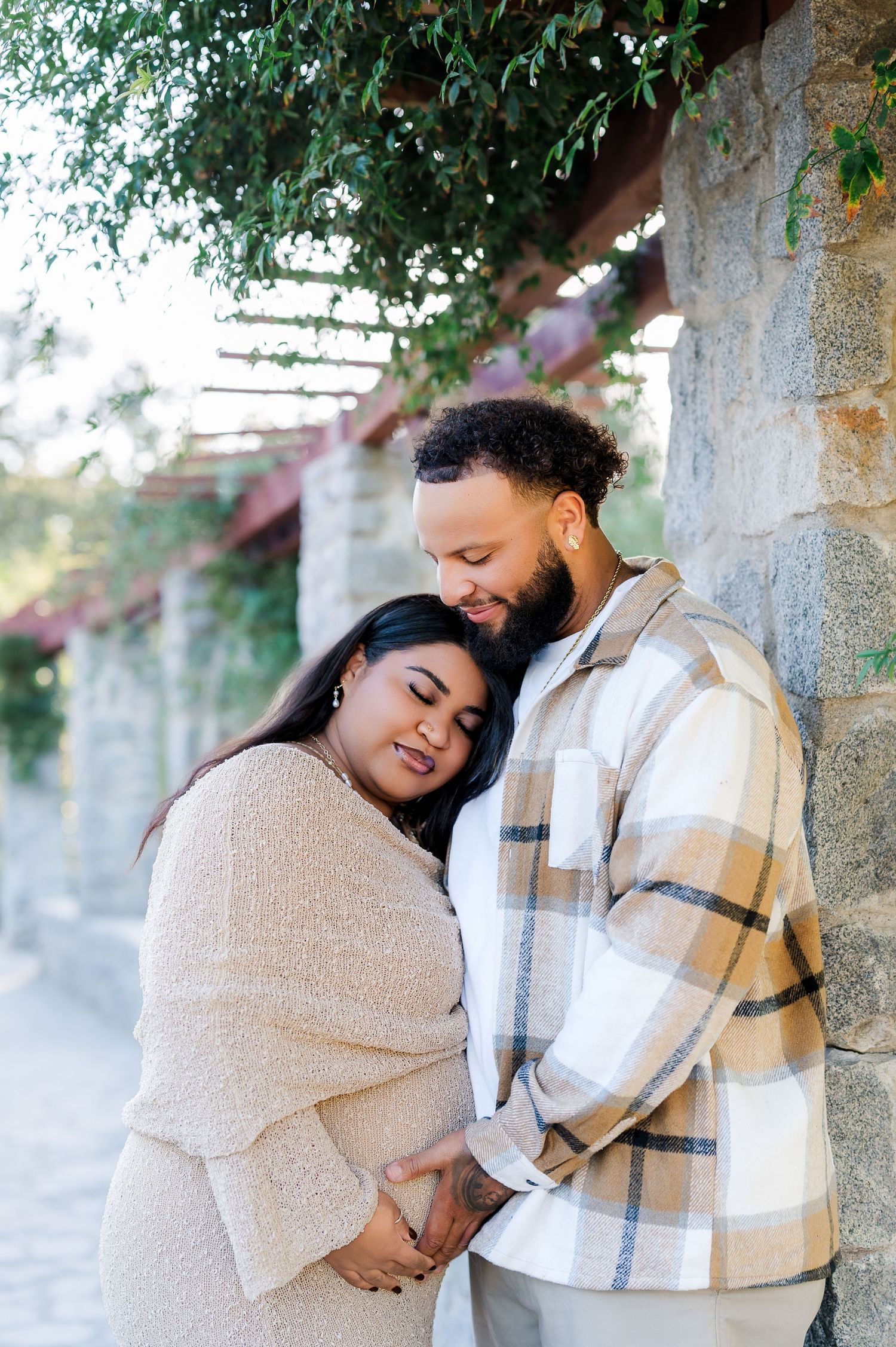A couple shares a tender moment leaning against a stone wall, the woman cradling her baby bump as her partner embraces her. WPA Rock Garden
