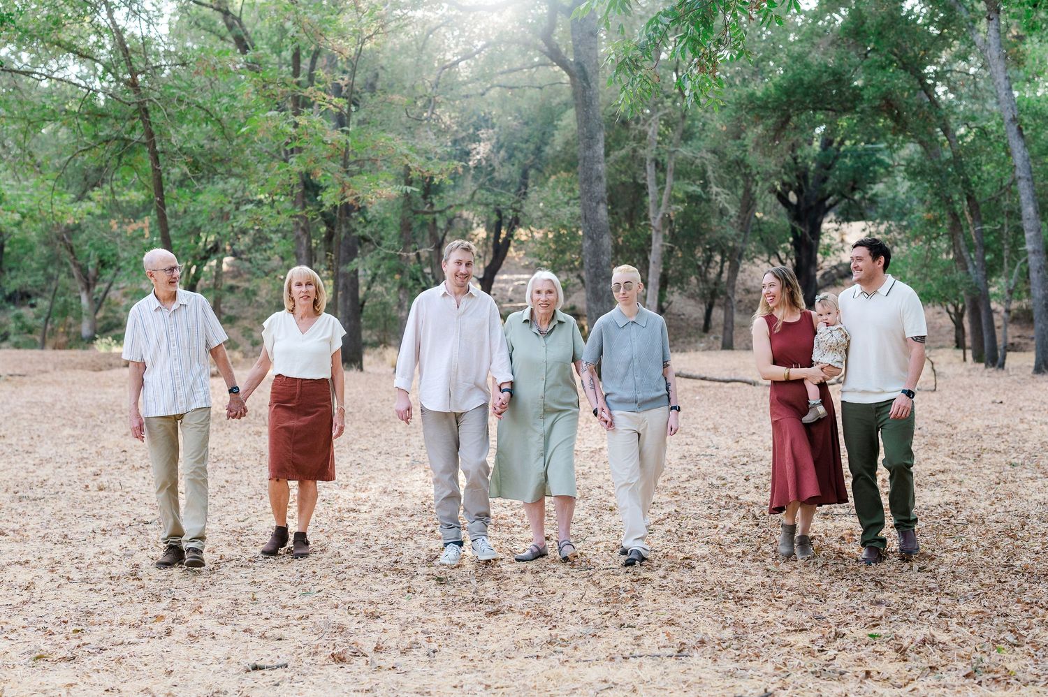 Multi-generational family holding hands walking together in a sunlit autumn park with fallen leaves.