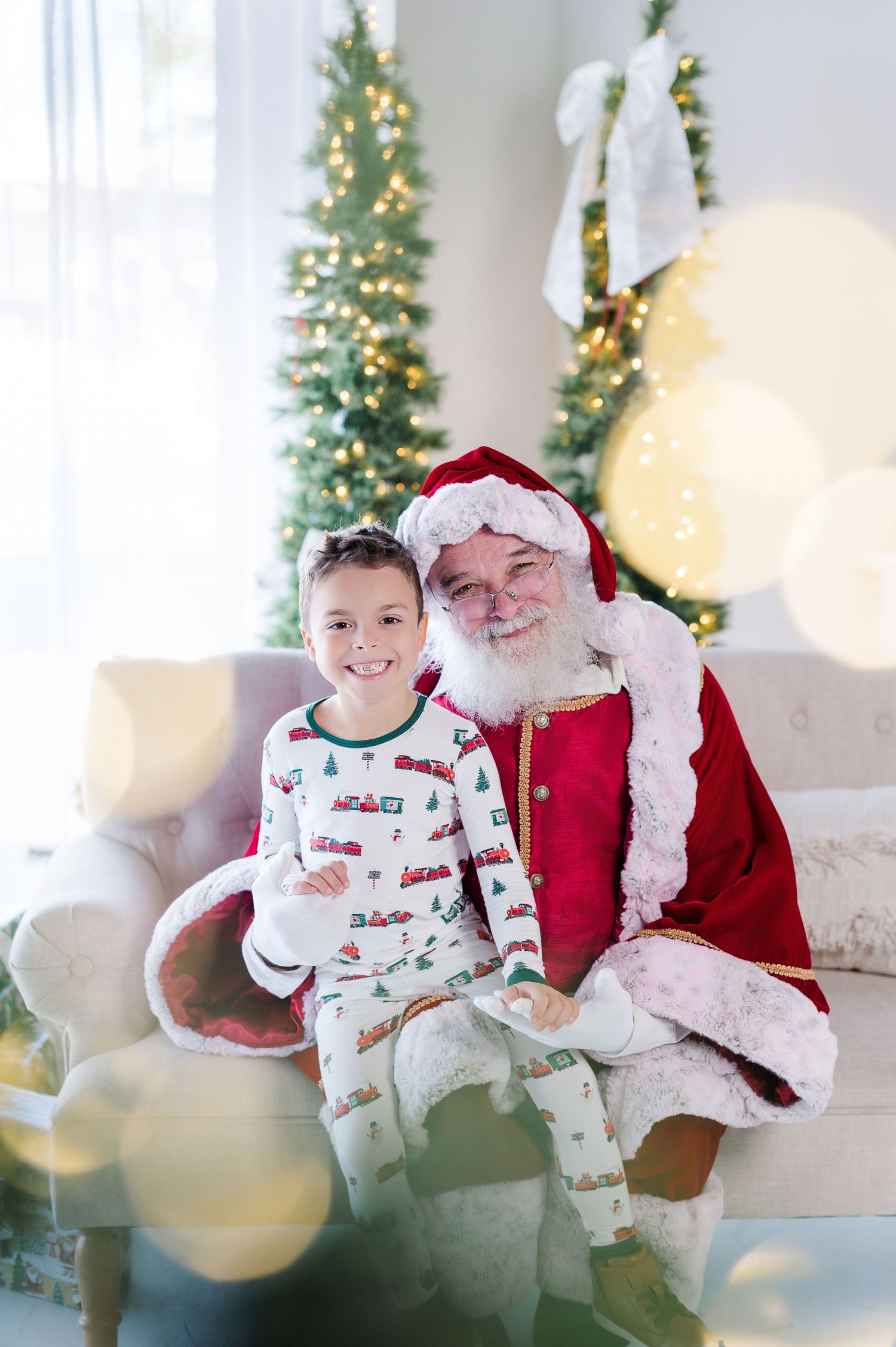 A smiling child in Christmas pajamas sits with Santa Claus on a couch beside a decorated Christmas tree.