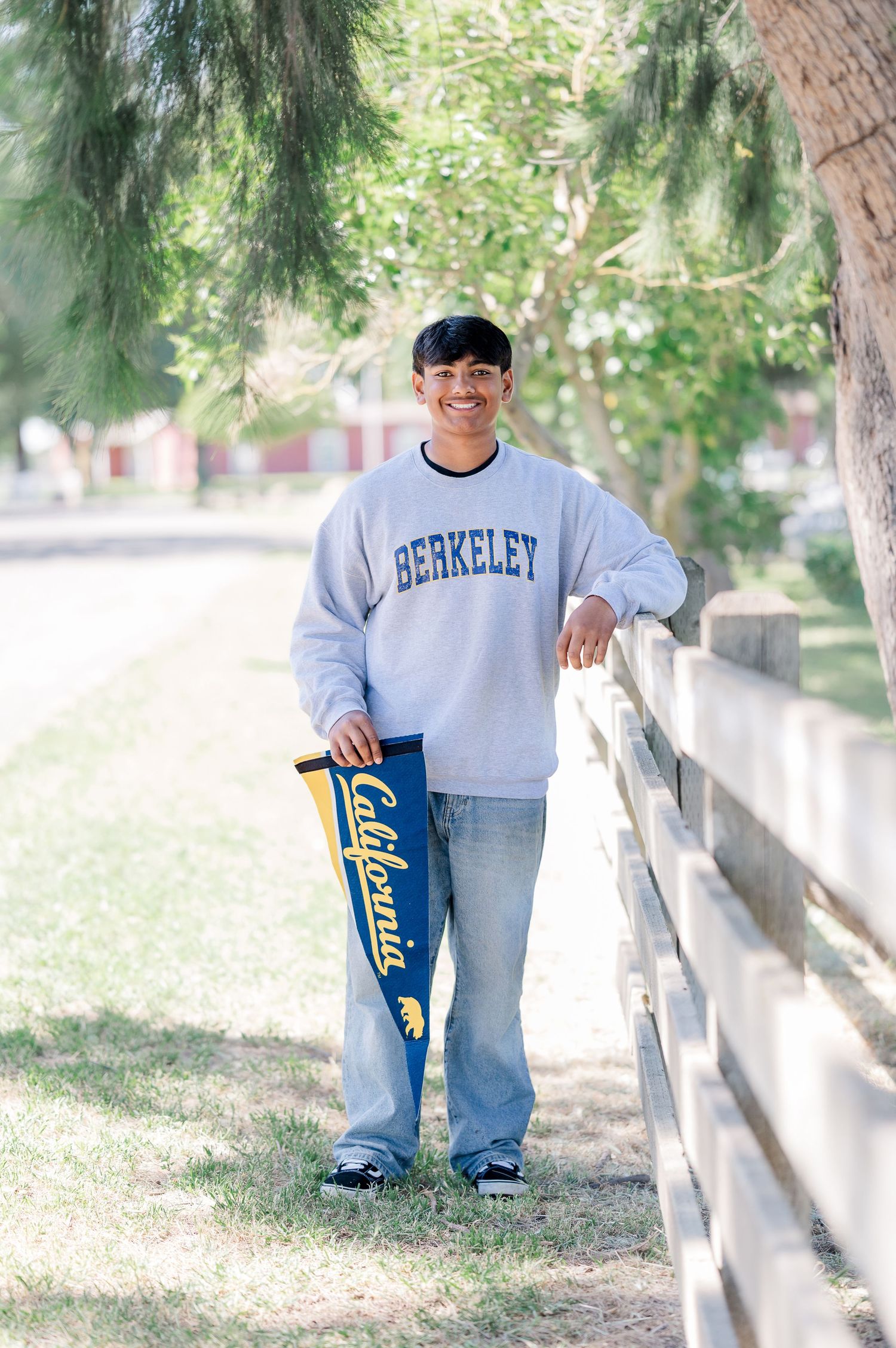 Young man in Berkeley sweatshirt holding California pennant, leaning on fence outdoors among trees.