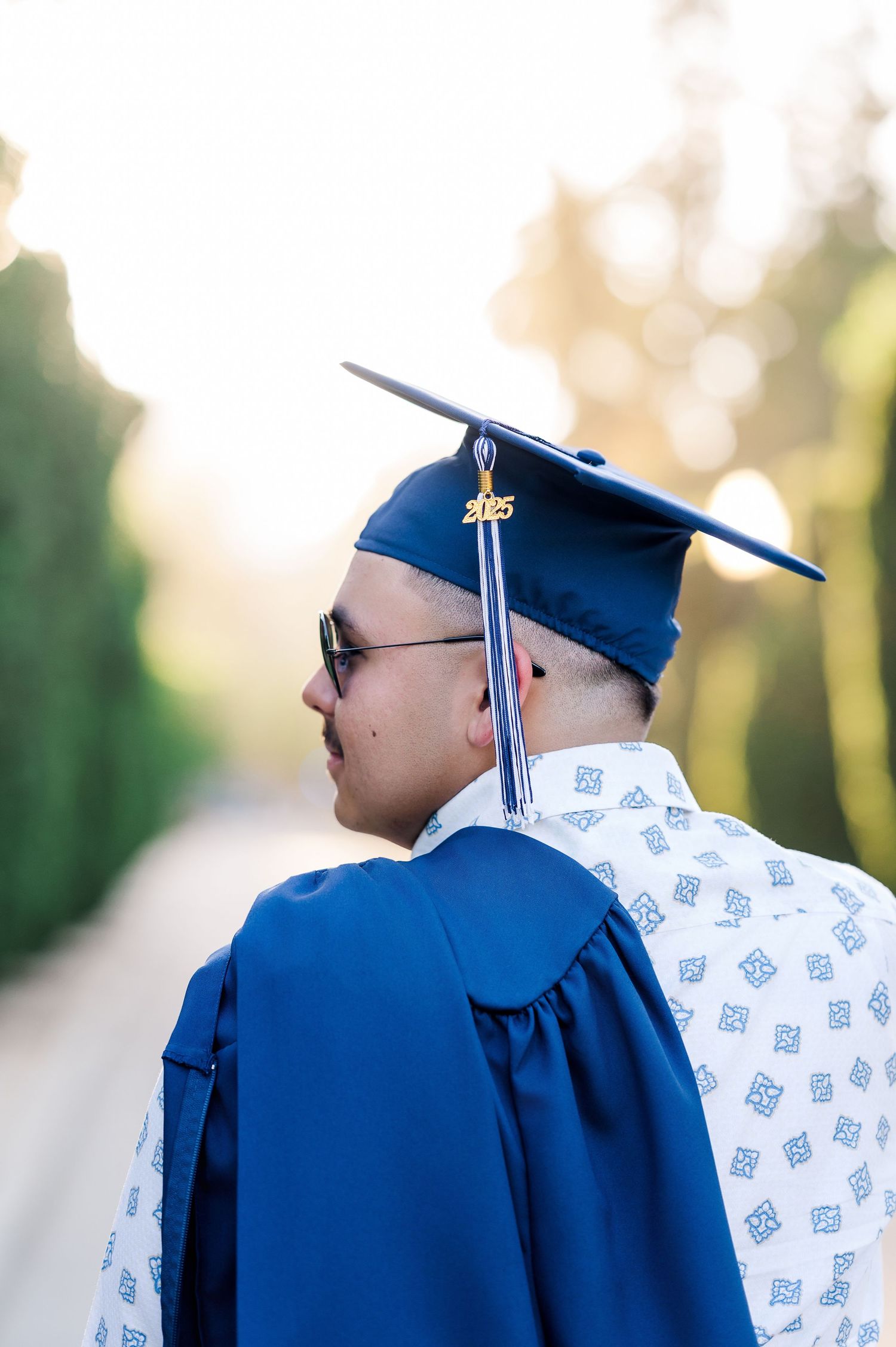 Graduate in blue cap and gown with 2025 tassel poses outdoors among lush greenery in warm sunlight.