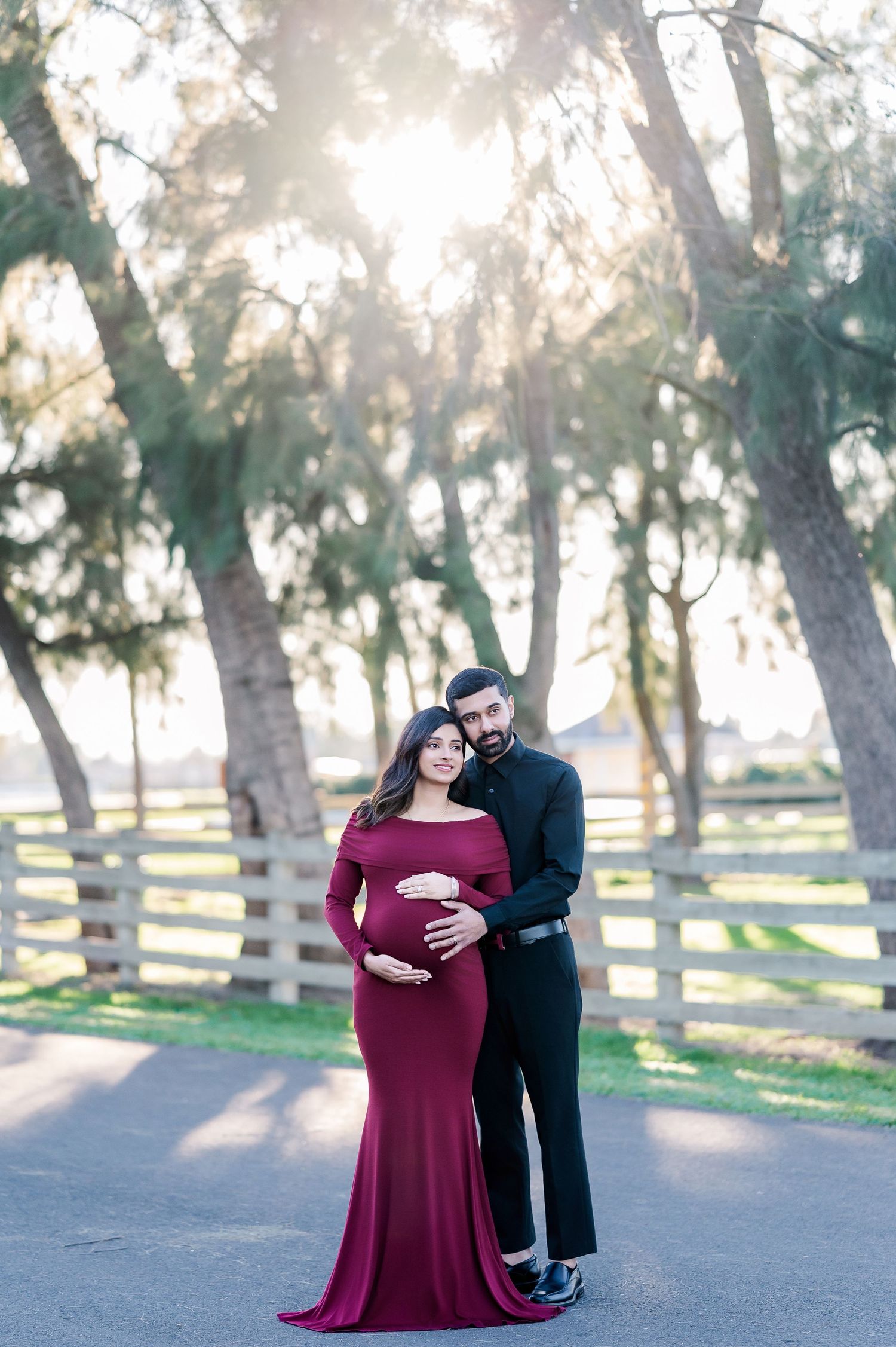 A romantic maternity photoshoot in a wooded park with a couple in burgundy dress and black attire at sunset.