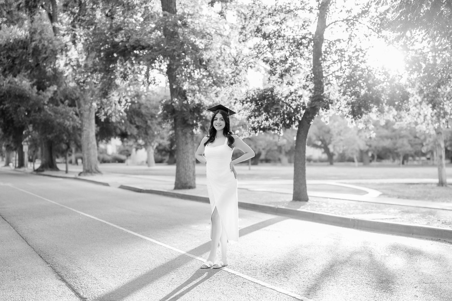 Black and white photo of graduate in white dress standing on tree-lined campus path.