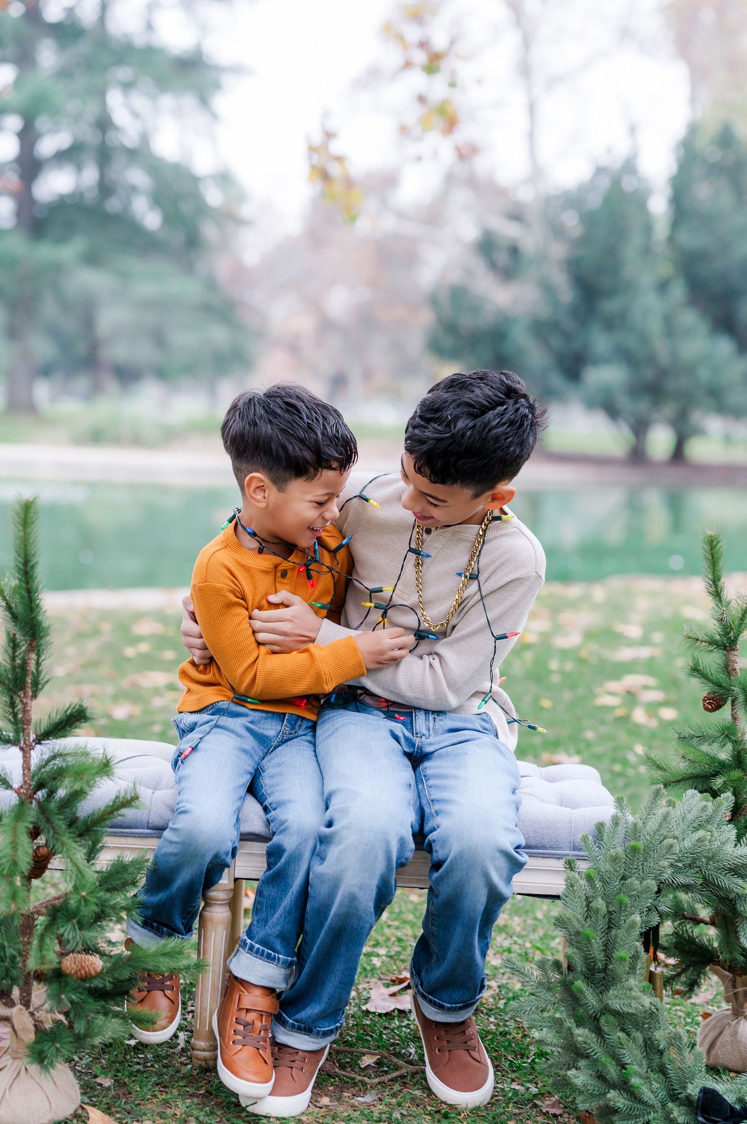 Two young children sitting together on a bench between evergreen trees in an autumn park setting.