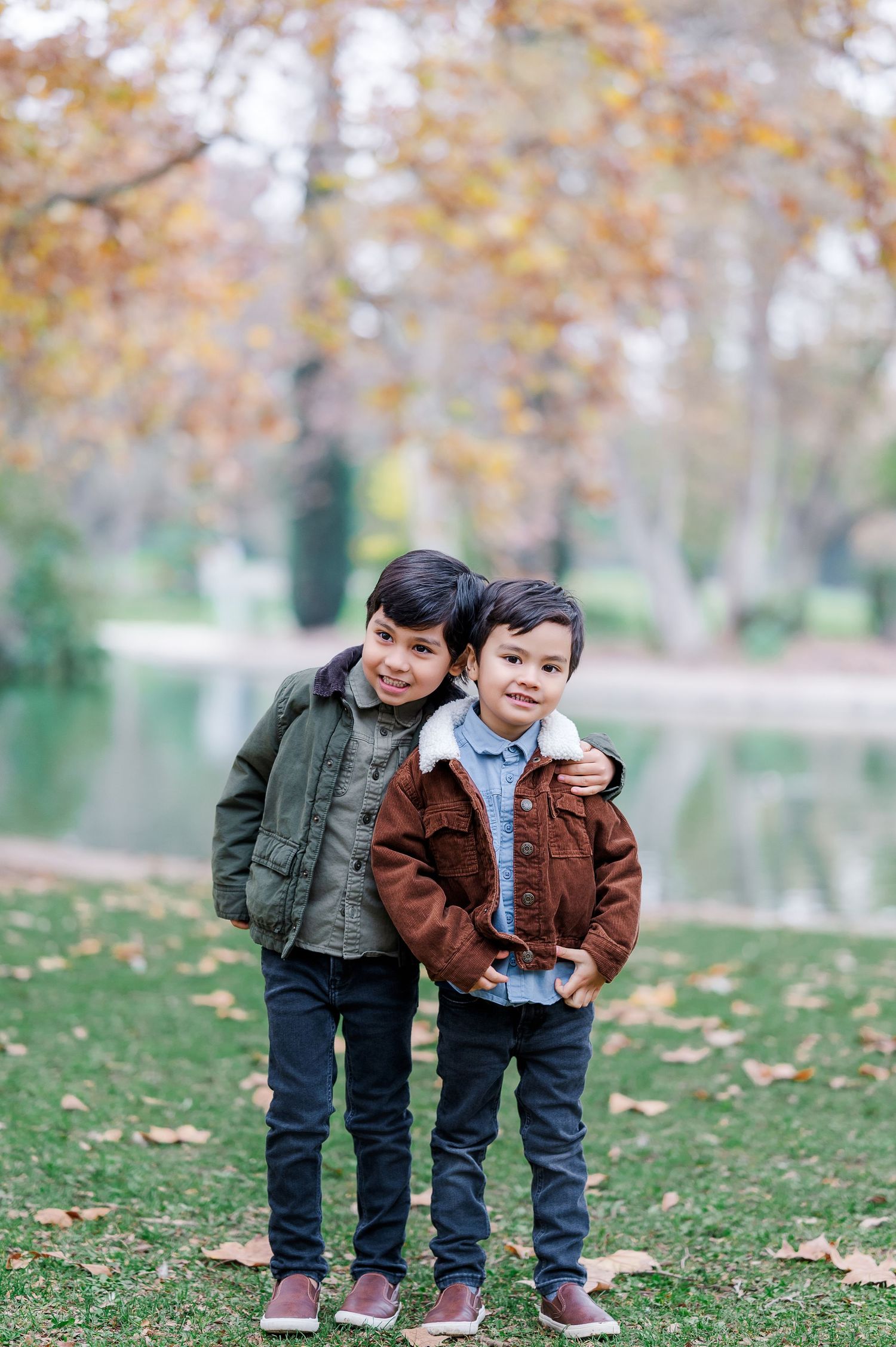 Two children in jackets play together on grass covered with autumn leaves near a pond.