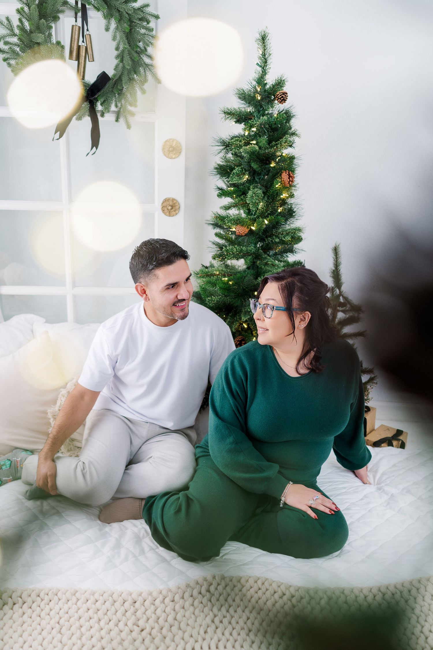 Couple in white and green outfits share tender moments while relaxing on white bedding with Christmas decor in background.