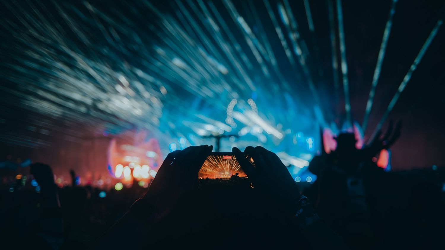 Silhouettes of concert crowd with raised hands against dramatic blue and orange stage lighting at a music festival.