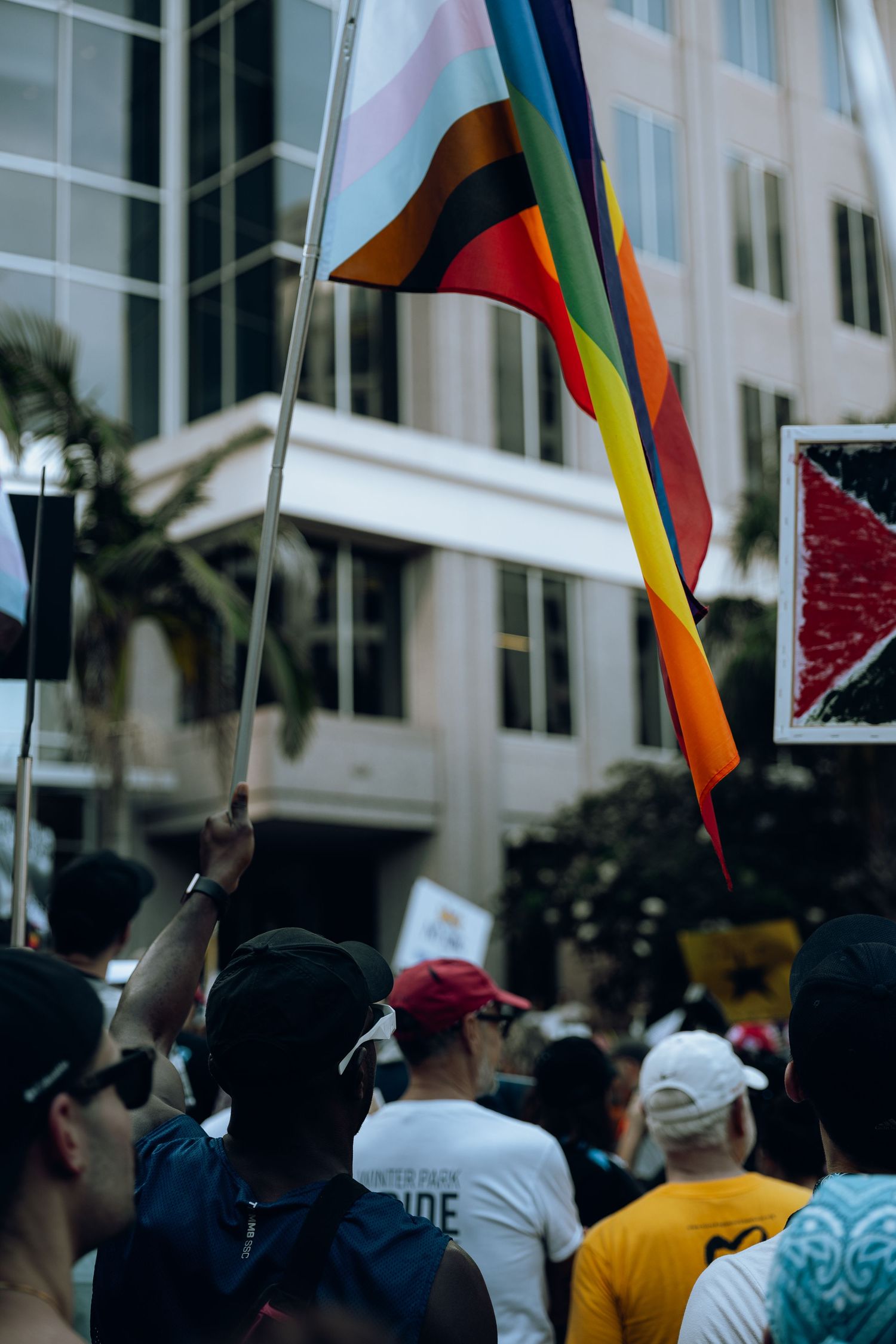 Colorful pride flag waves above crowd of protesters at urban demonstration.