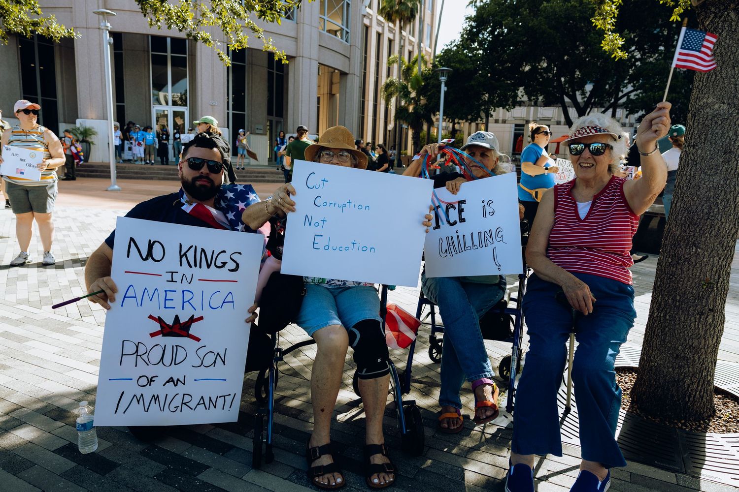 Protesters holding signs opposing monarchy and tyranny gather on a sunny sidewalk with an American flag visible.