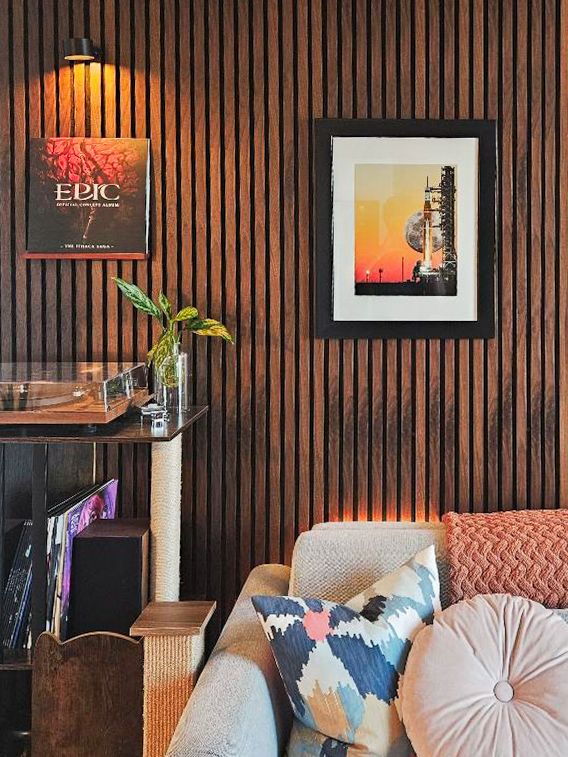 Cozy living room corner with slatted wood wall, framed rocket art, EPIC poster, and colorful accent pillows on sofa.