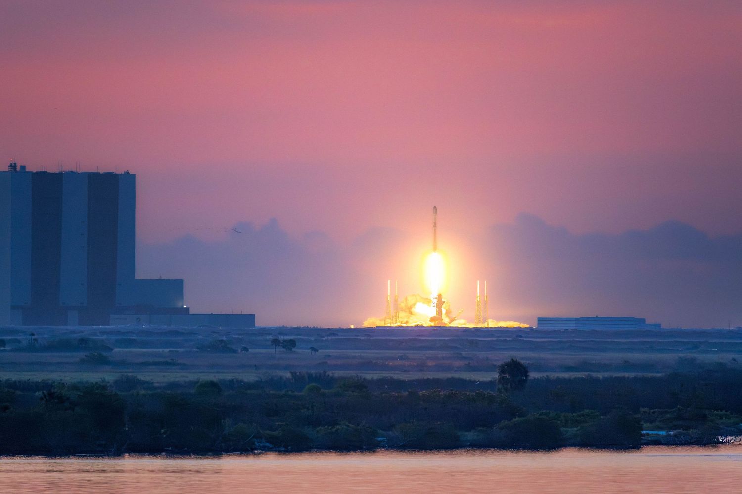 SpaceX rocket launch at sunset with vibrant pink sky over Kennedy Space Center launch pad.