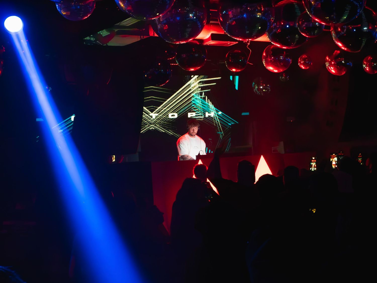 A bright blue spotlight beam illuminates a dark nightclub with red lighting and electronic music equipment visible.