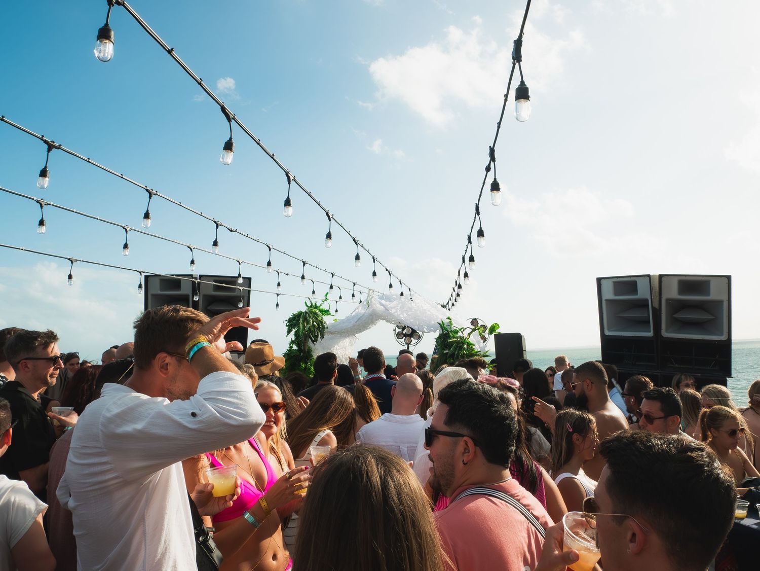 A crowded outdoor party scene with string lights overhead and people dancing at a daytime rooftop event.