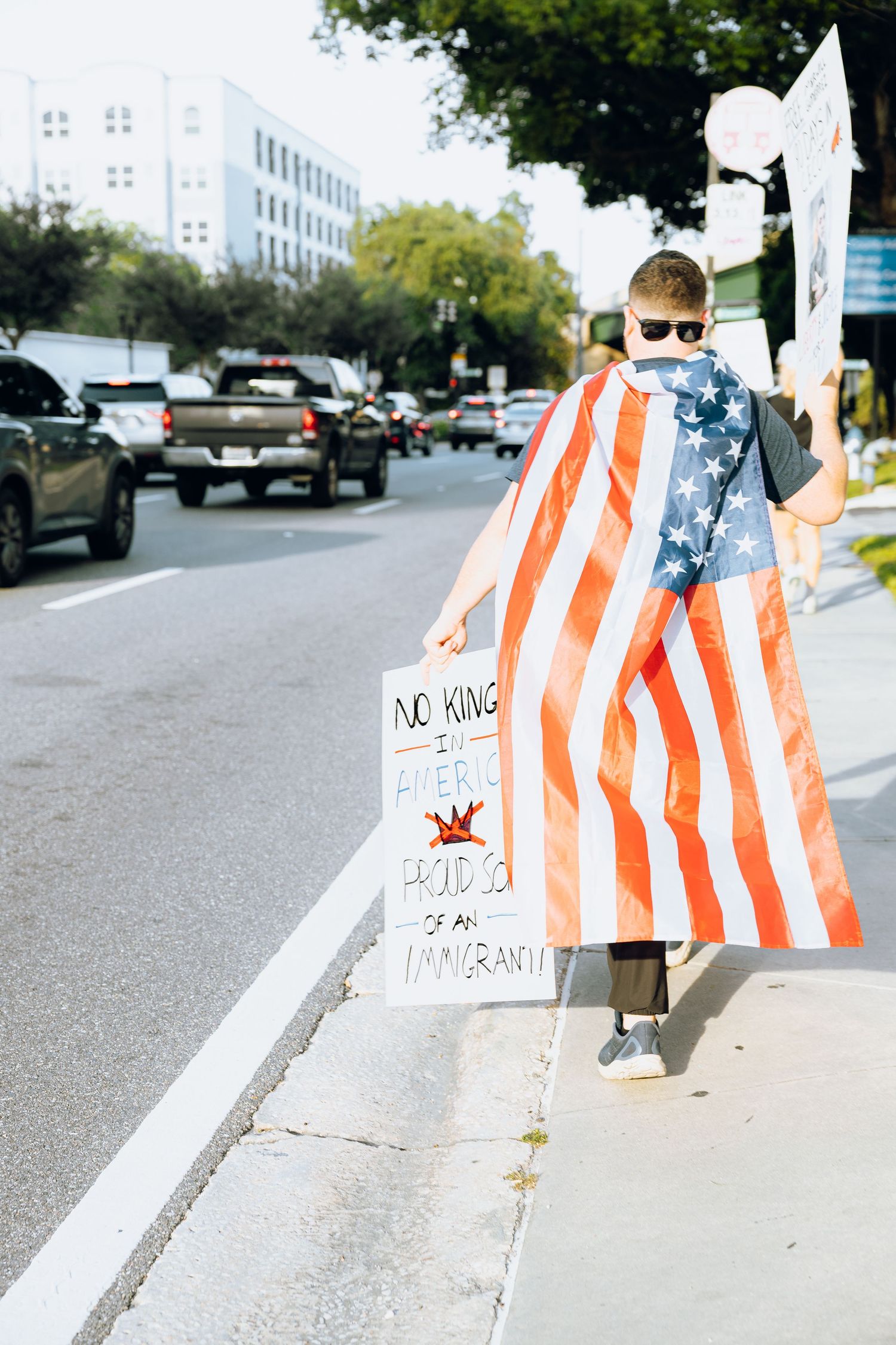 Person wearing American flag cape carrying protest sign while walking down urban sidewalk.