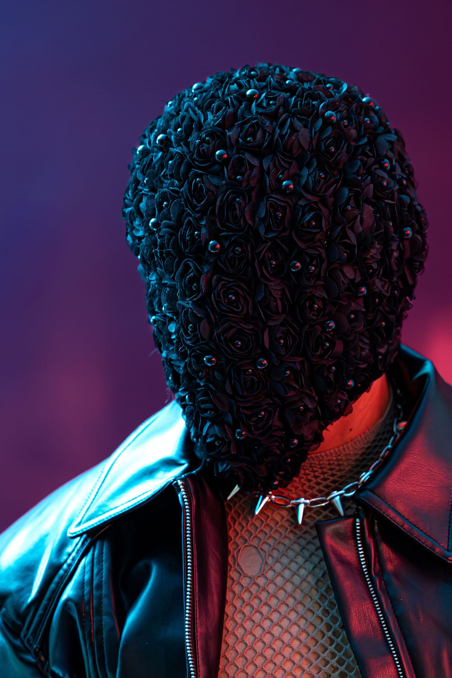 A figure wearing a black rose-covered mask, leather jacket, mesh top, and spiked choker under dramatic neon lighting.