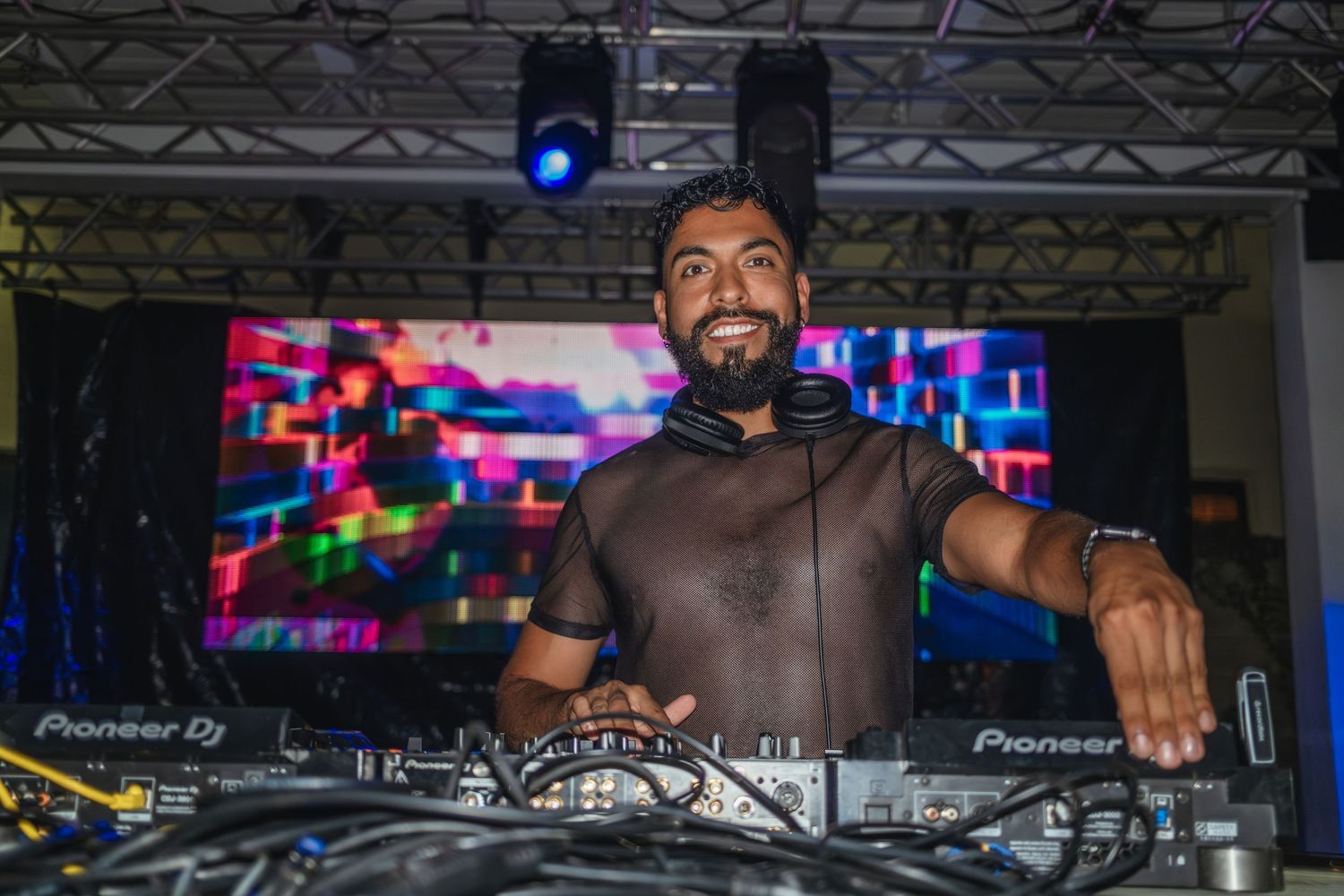 DJ performs behind turntables at a nightclub with colorful lights and electronic equipment in the background.
