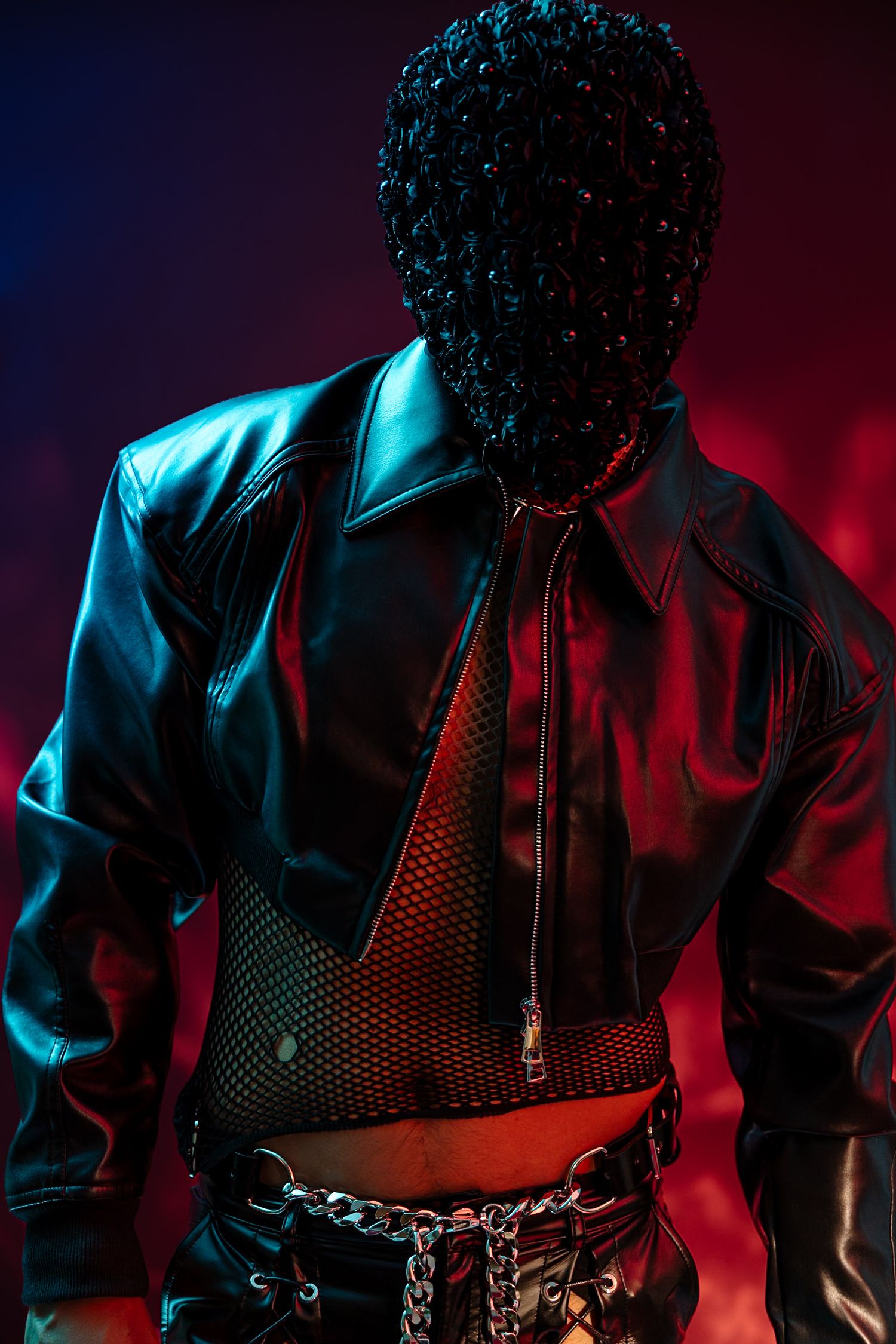 Person in black leather jacket and mesh top with chain belt, wearing a textured black mask under dramatic neon lighting.