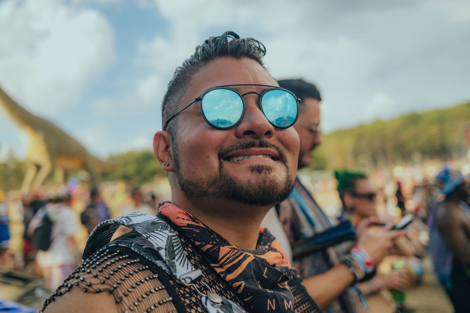 Festival goers wearing sunglasses enjoy a sunny outdoor music event with a crowd visible in the background.