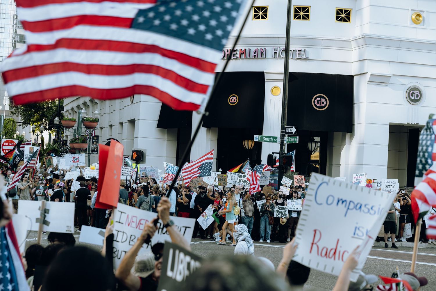 Protesters gather at a rally holding signs and American flags in front of retail storefronts.