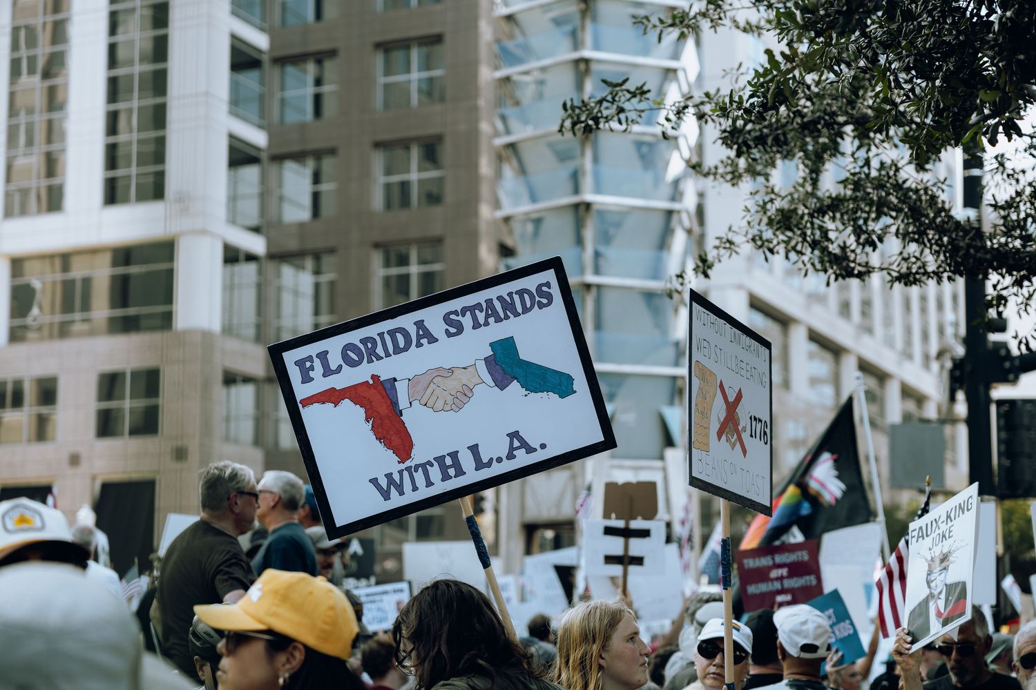 Protesters at a rally hold up signs including one that reads 'Florida stands with L.A.' among city buildings.