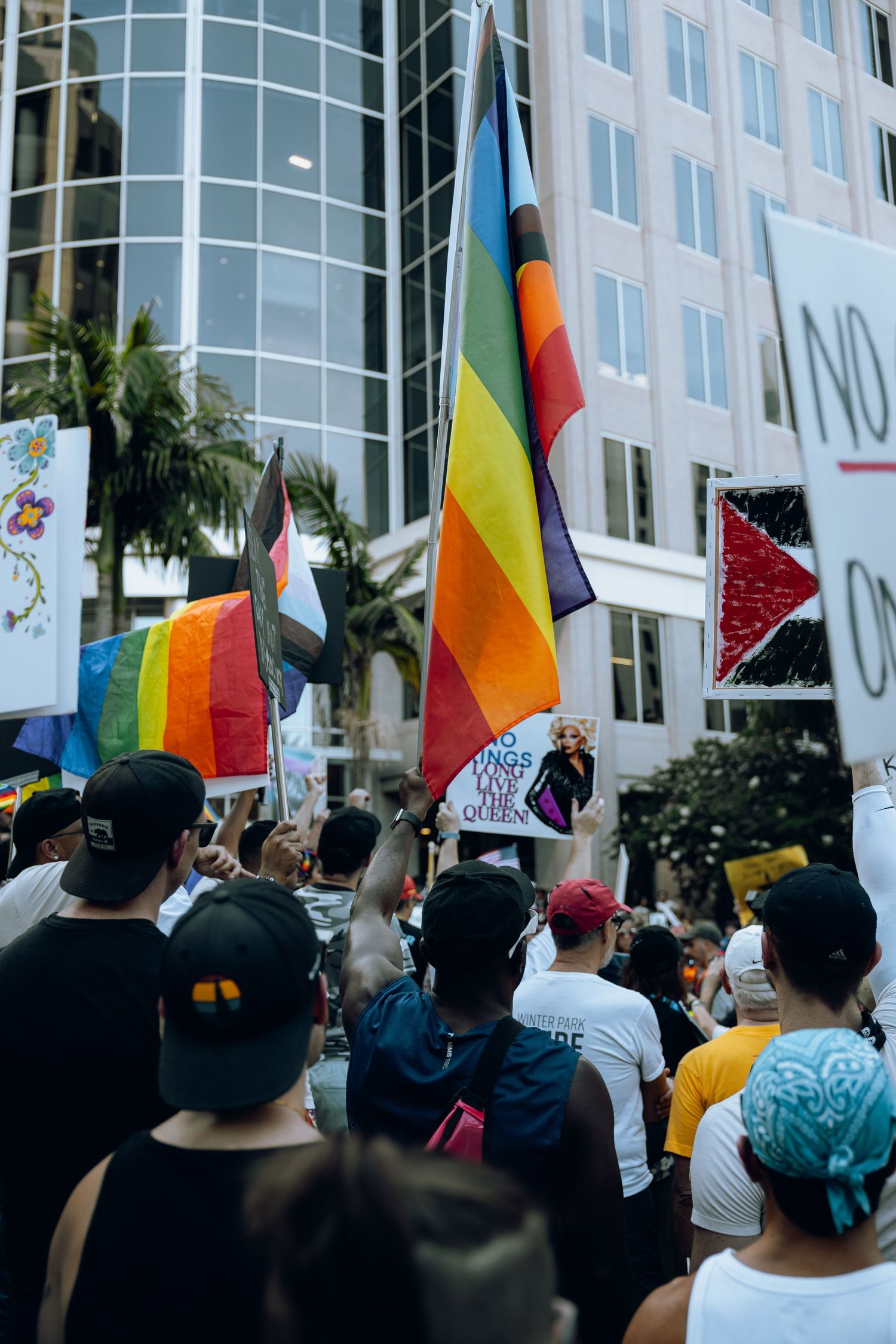 Protesters hold rainbow pride flags and gather outside a modern office building during a demonstration.