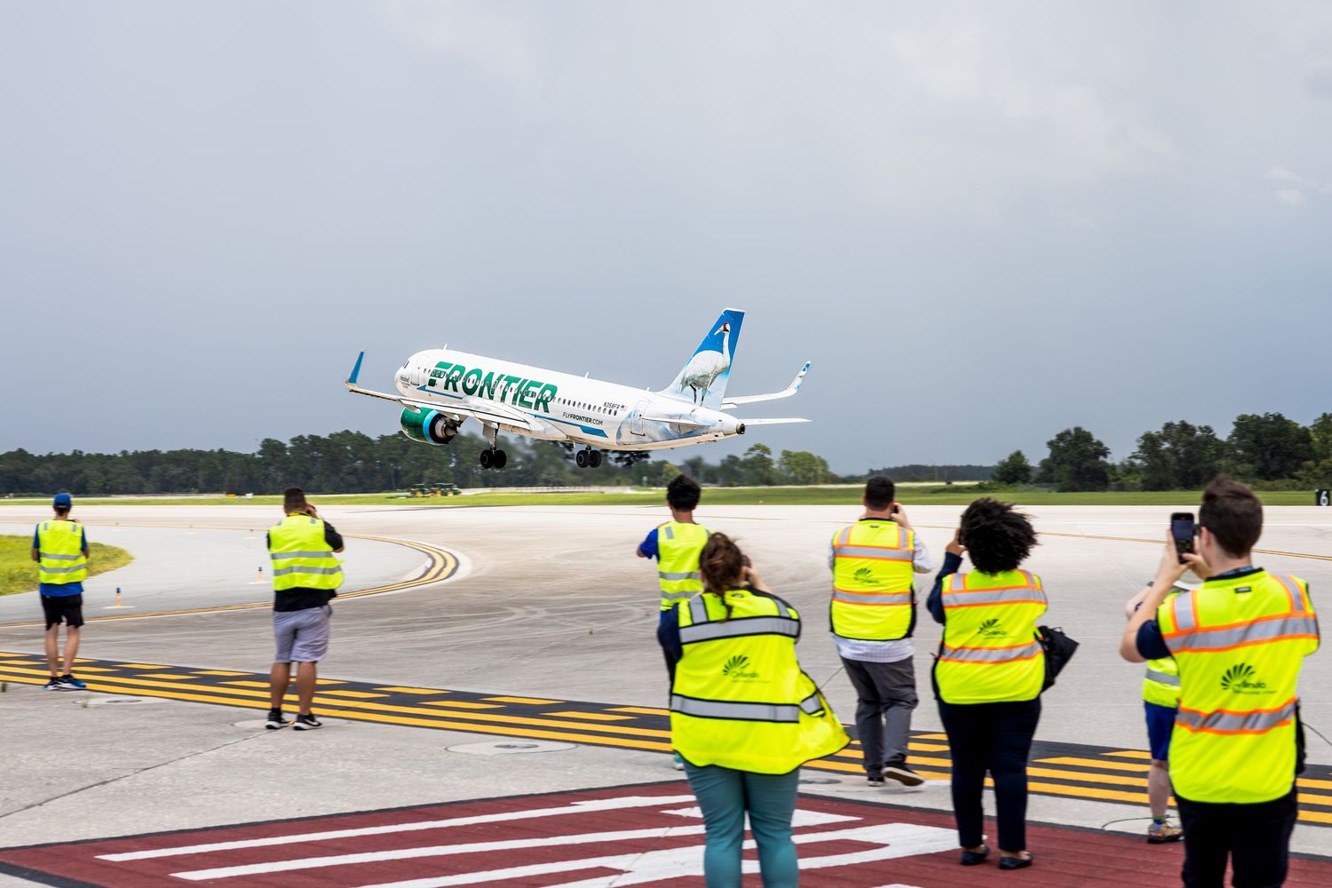 Frontier Airlines plane taking off at airport with ground crew in yellow vests watching from tarmac.