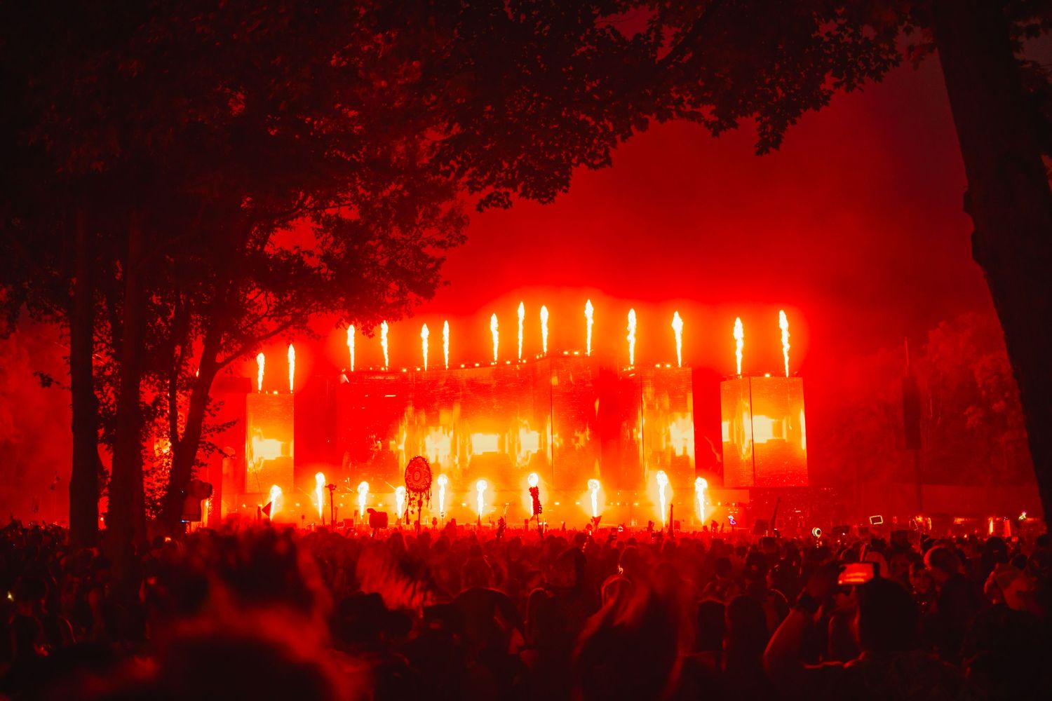 Massive outdoor music festival stage lit with intense red and orange lights amid trees at night.