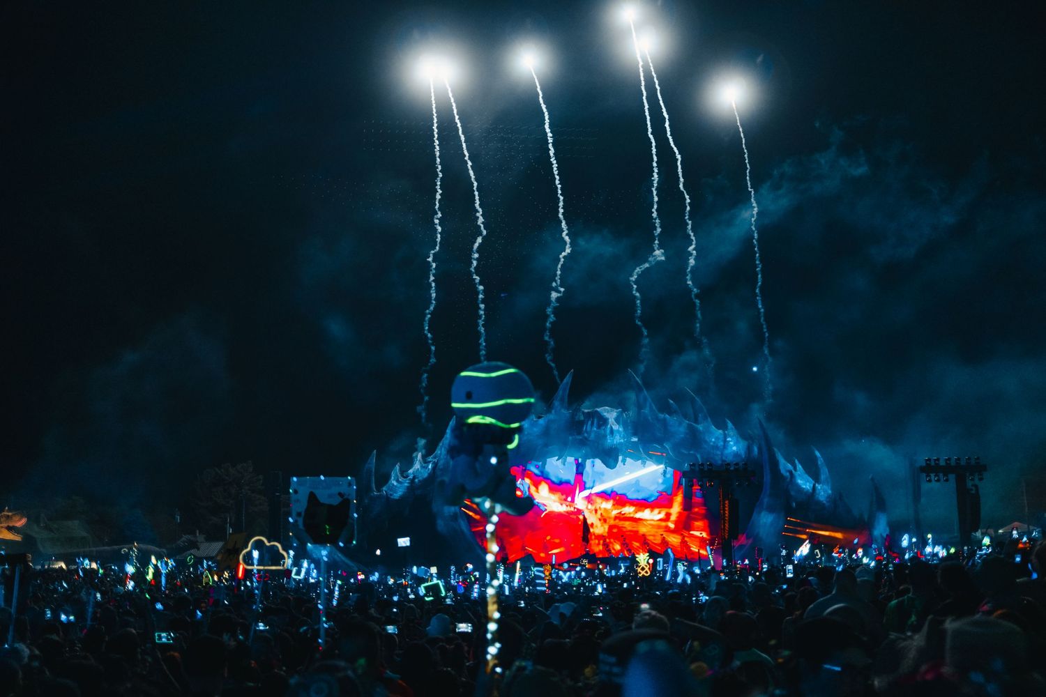 Lightning bolts illuminate a dark night sky above a festival stage with bright lights and a large crowd.