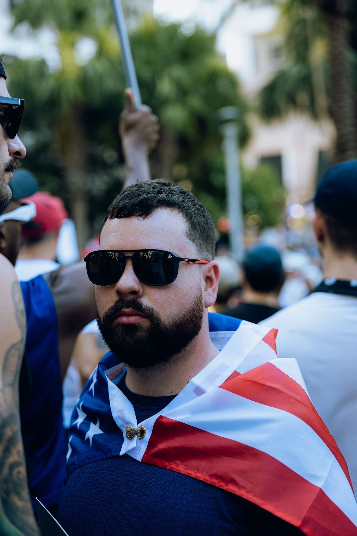 Person wearing sunglasses and an American flag draped as a cape during a protest gathering.