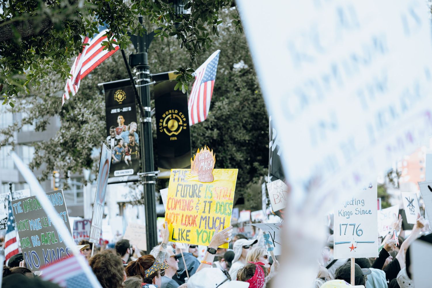 Protest crowd gathering with American flags and signs visible through blurred foreground.