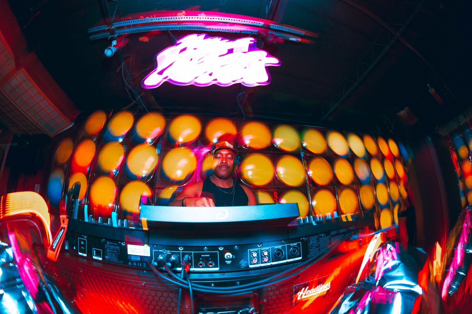 DJ performing at a vibrant nightclub with colorful bokeh lights and neon signs in a fisheye lens shot.