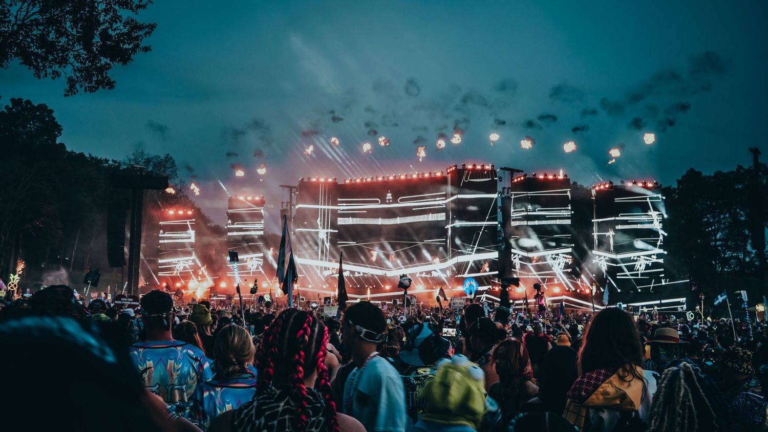 Concert crowd silhouettes against dark sky with stage lights and equipment visible in outdoor venue setting.