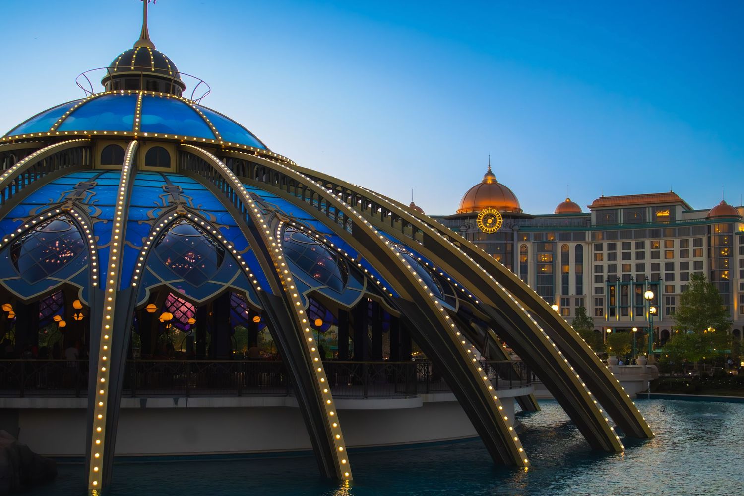 Illuminated arched pavilion with decorative lights sits by a lake near luxury buildings under blue sky.