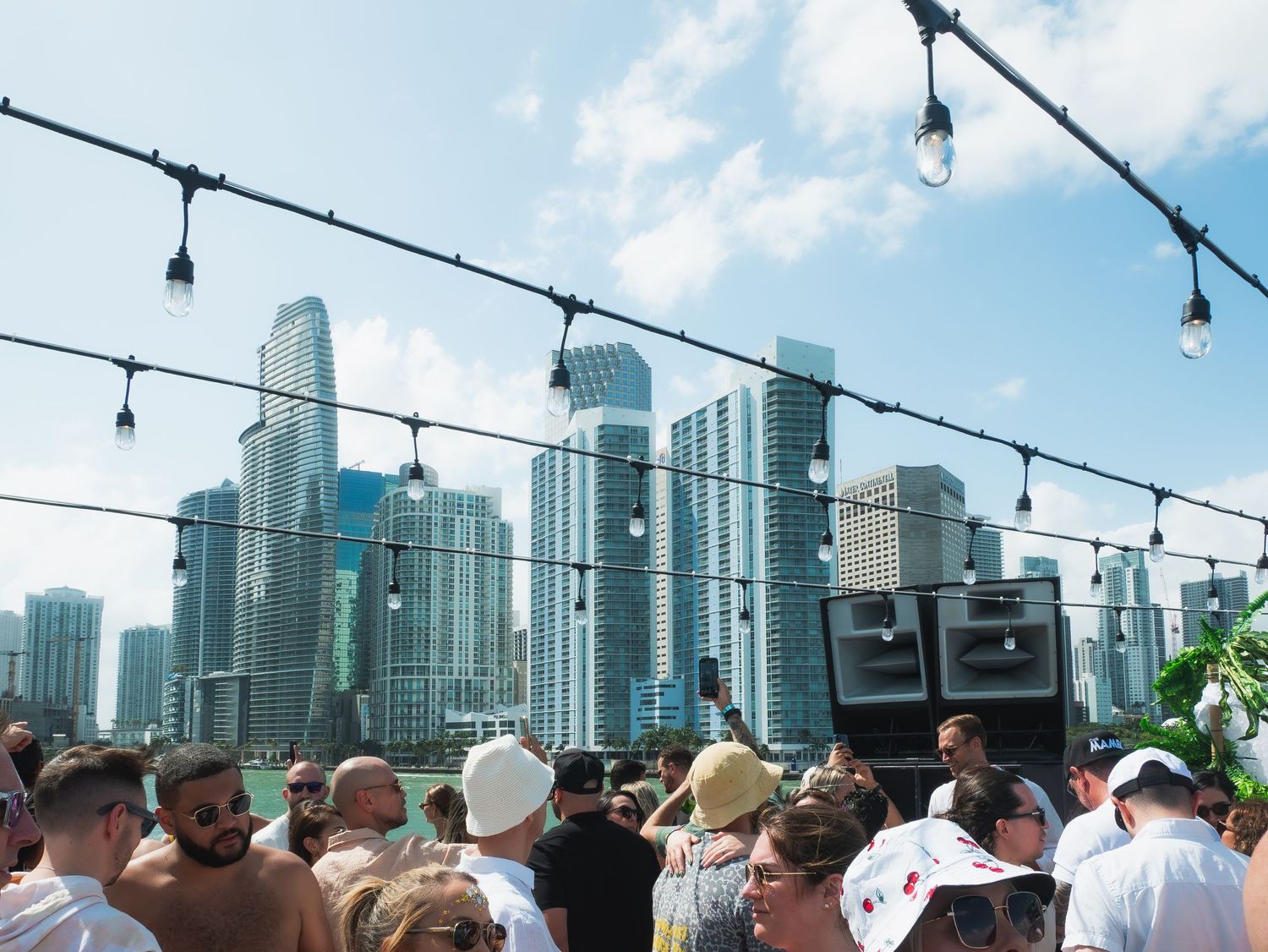 Rooftop party scene with string lights overlooking downtown Miami skyline on a sunny afternoon.