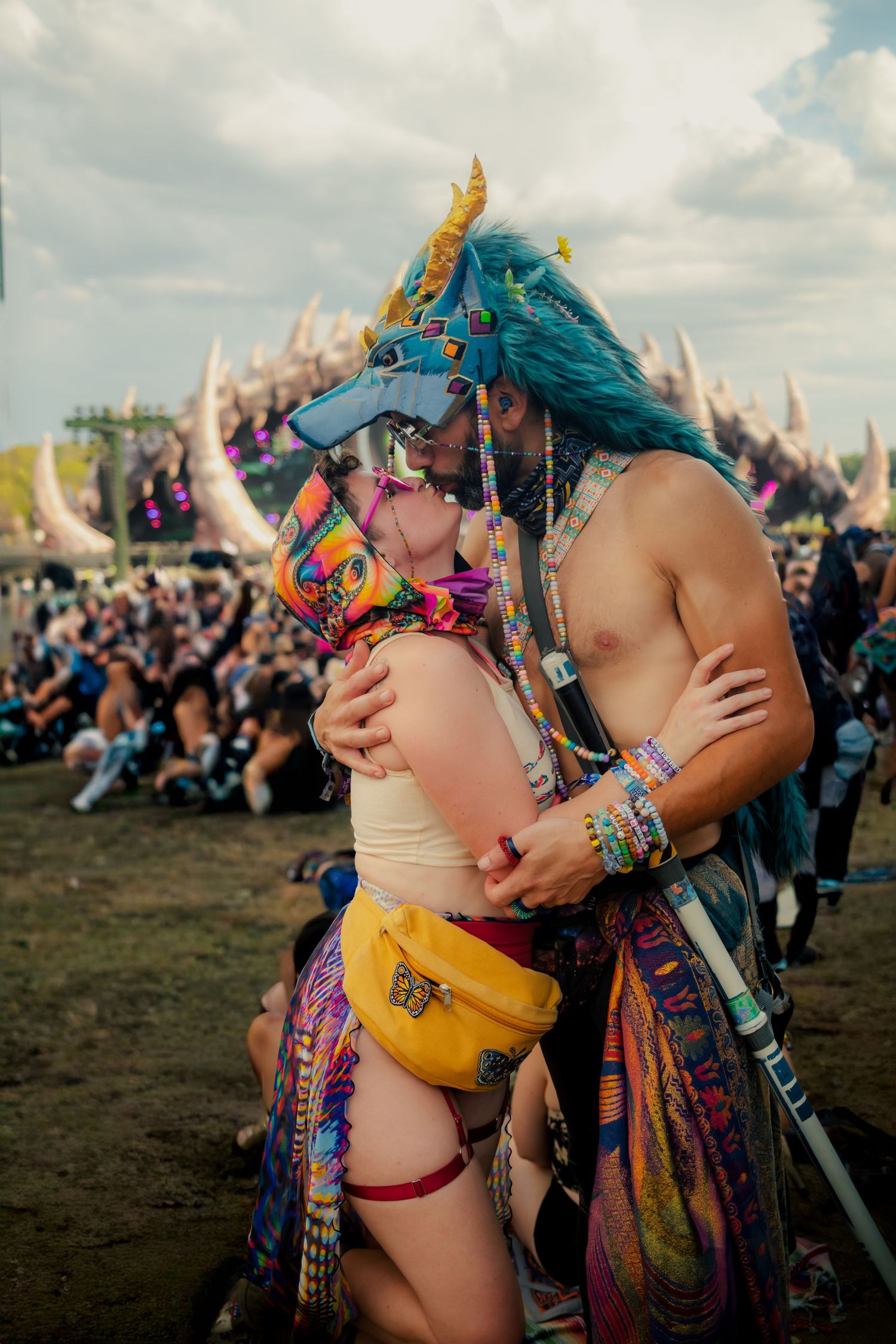 Festival goers in colorful festival attire embrace at an outdoor music event with cloudy skies in the background.