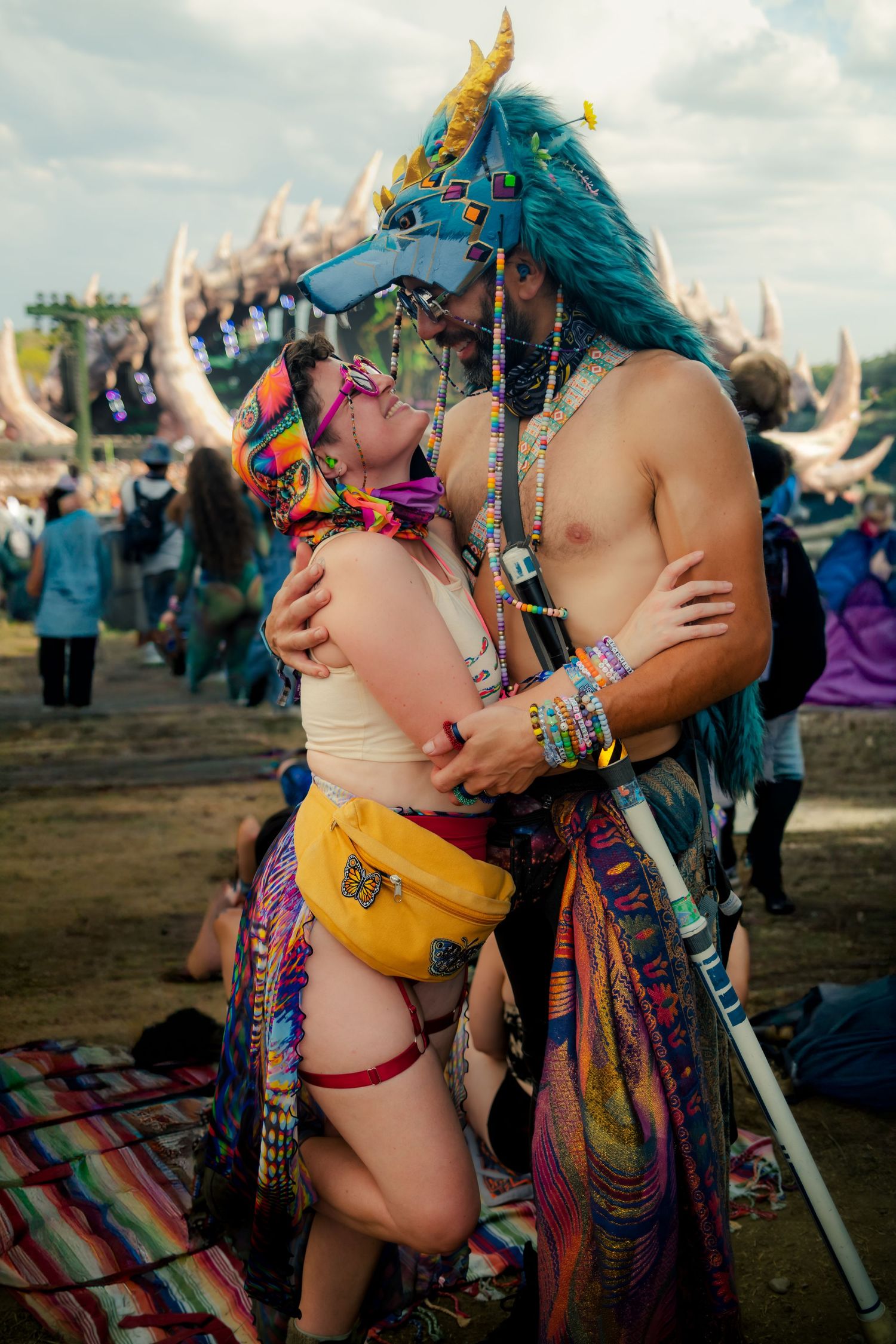 Festival attendees in colorful outfits embrace at an outdoor music event with tents and crowds visible in background.