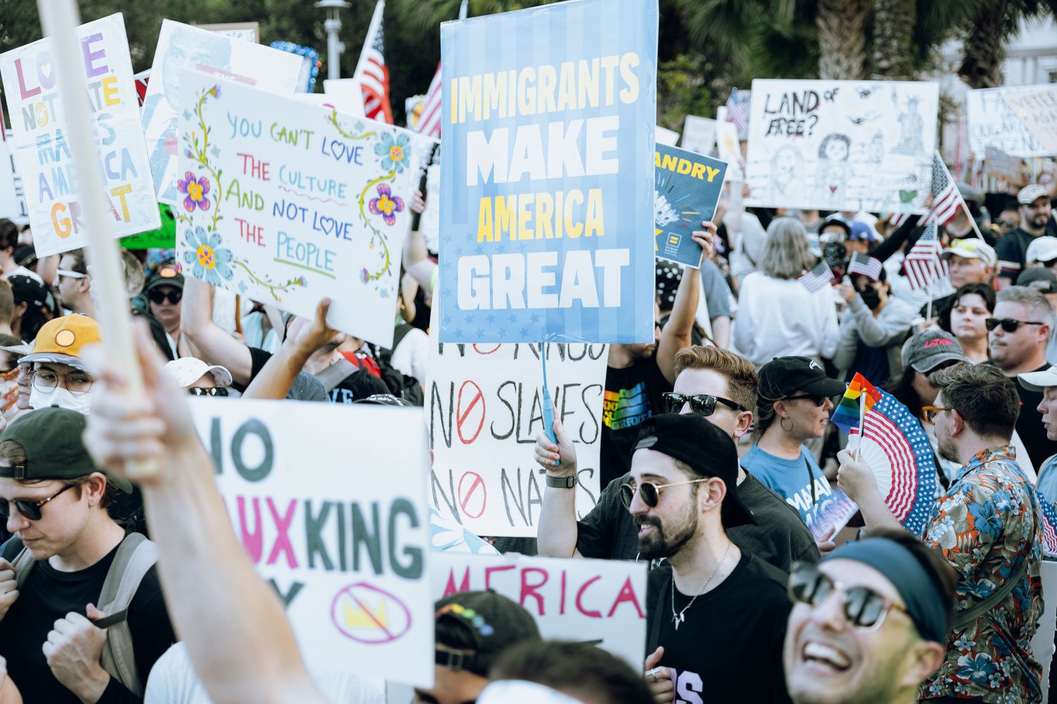 Large crowd of demonstrators holding protest signs at a daytime rally with colorful banners visible.