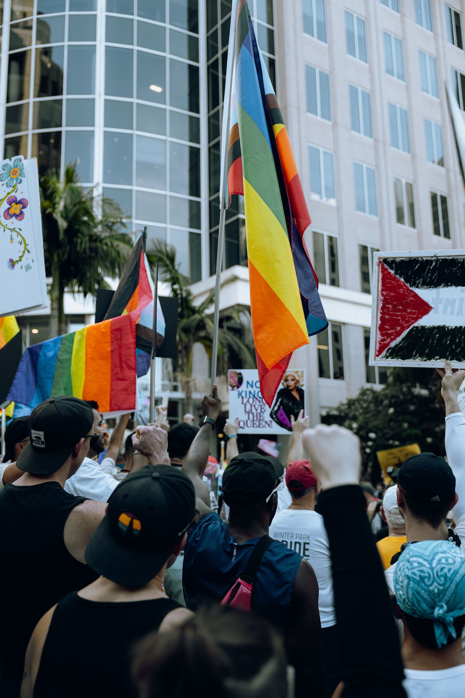Pride flags and protesters gathered outside a glass office building with palm trees.