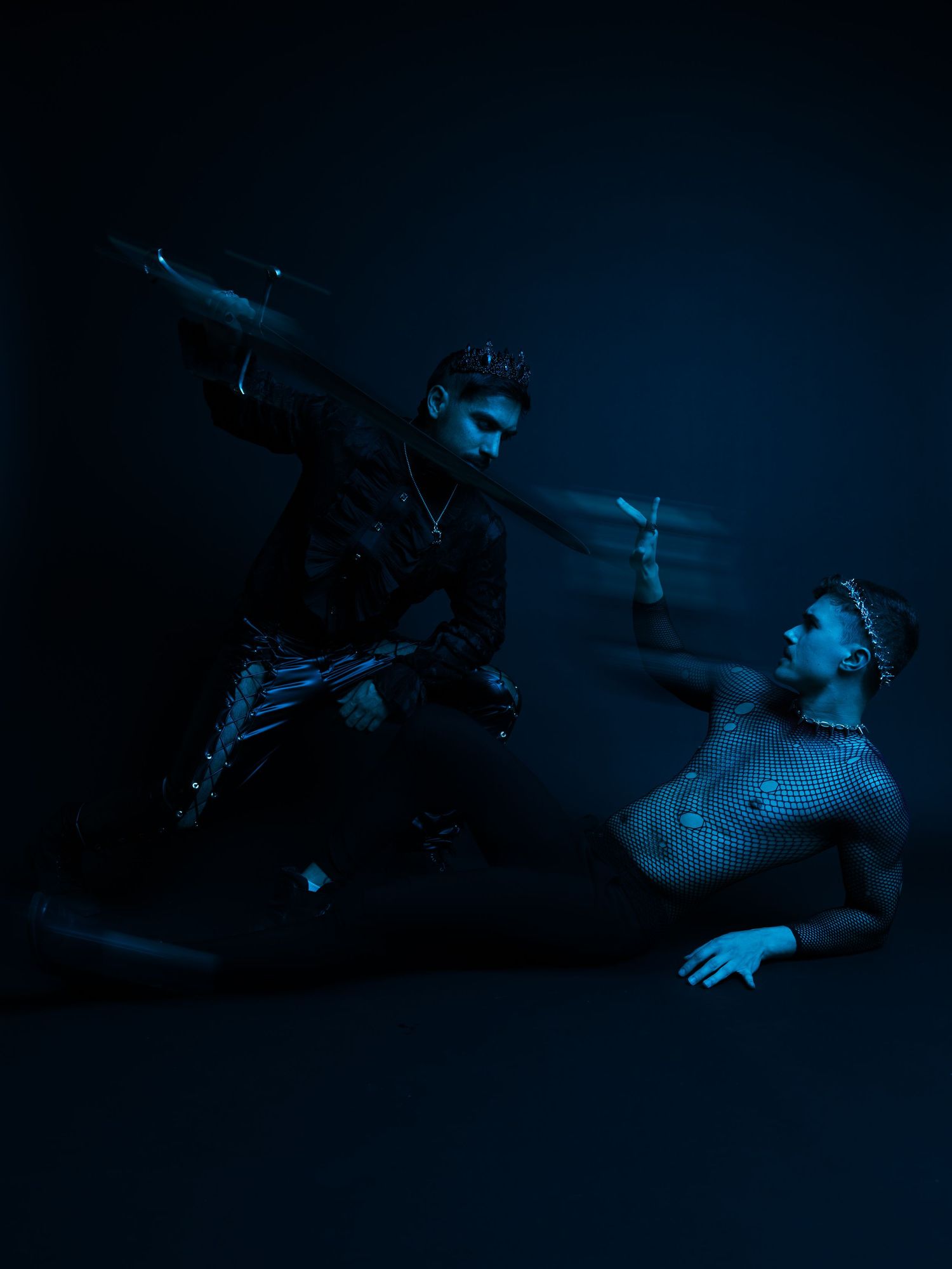Two dancers perform dramatically under blue lighting, one seated while the other poses dynamically in dark avant-garde fashion.