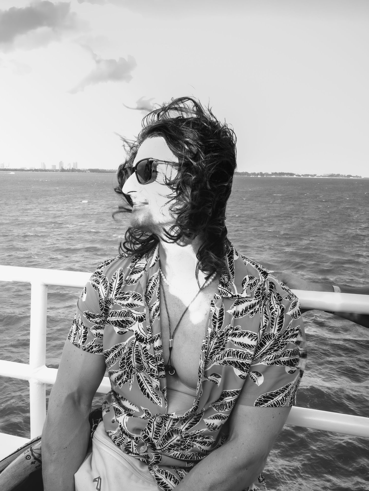 Black and white photo showing someone in sunglasses and a tropical print shirt on a boat deck with the ocean behind them.