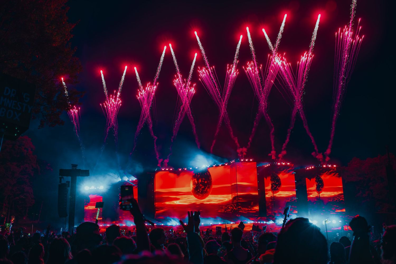 Pink fireworks explode above a concert stage with red lighting and a large crowd silhouetted against the dramatic display.