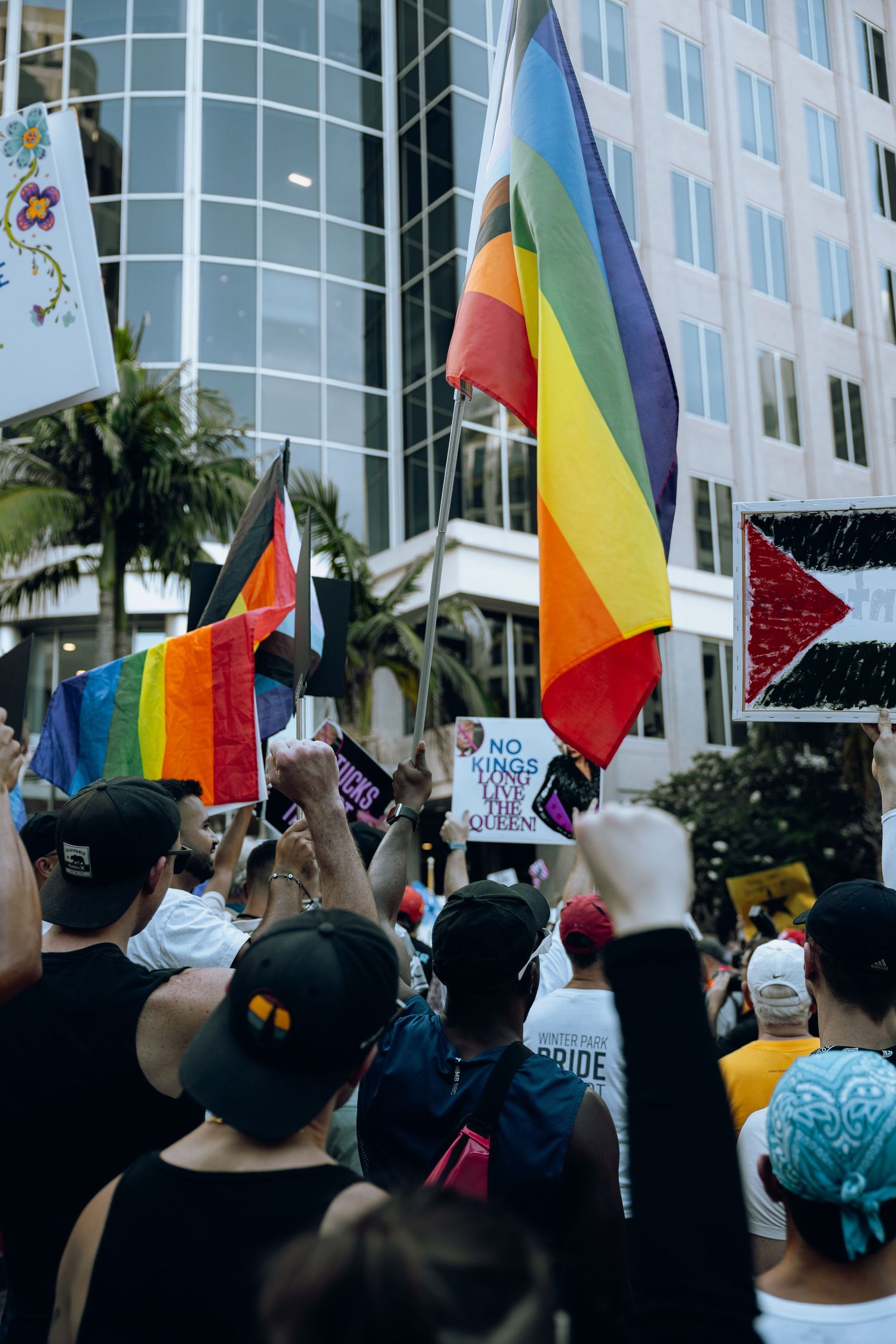 Crowd gathering with rainbow pride flags and protest signs in front of modern office building with palm trees.