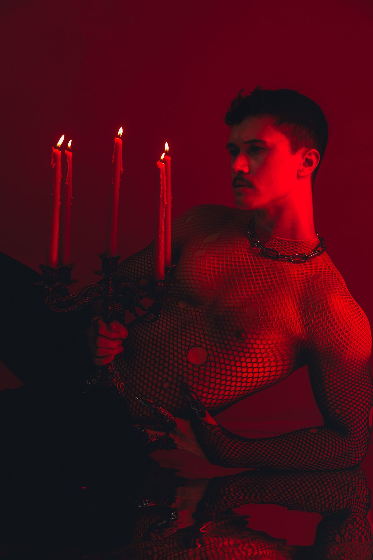 Shirtless man in mesh top holding candelabra with lit candles in dramatic red lighting.