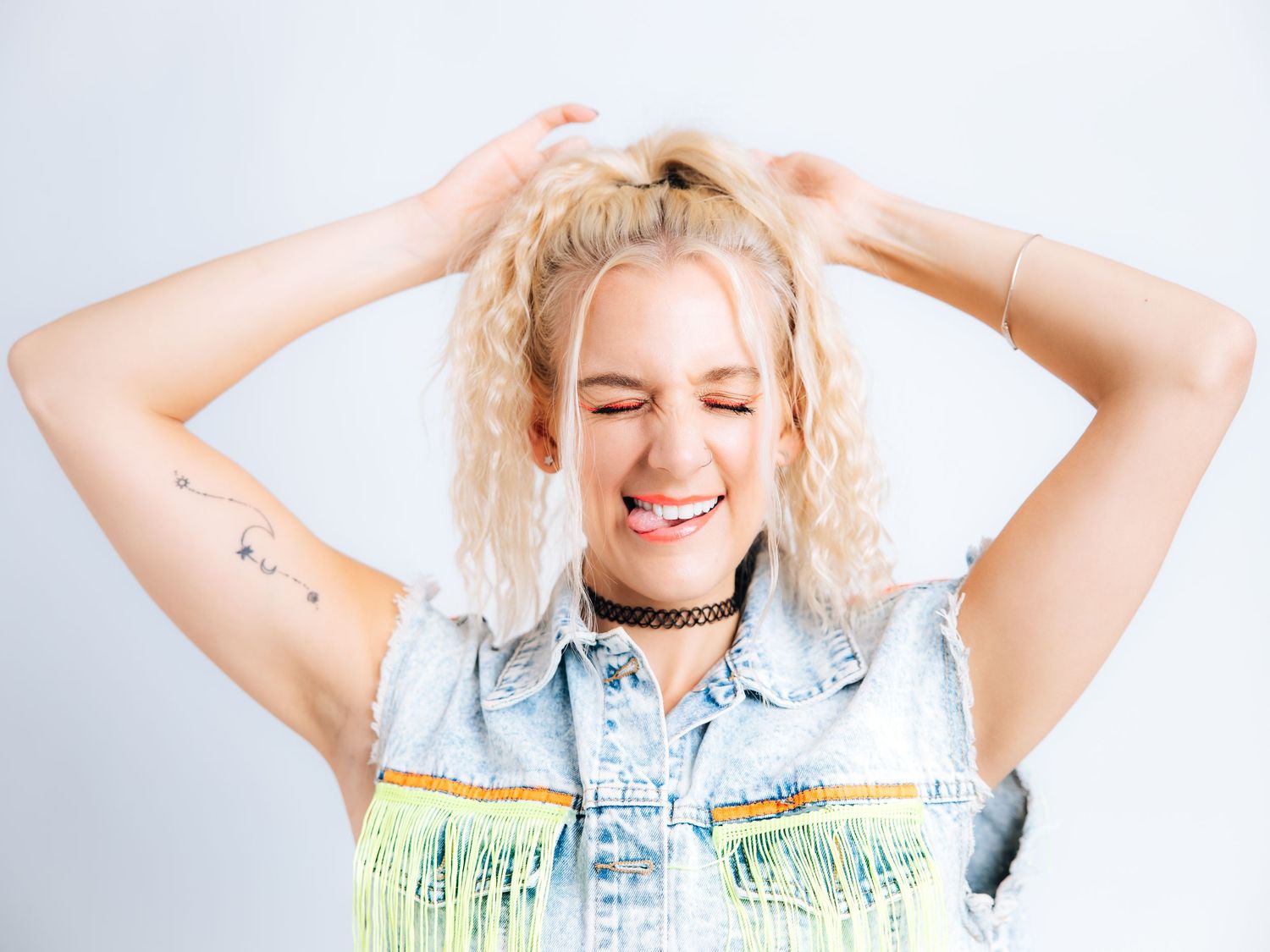 Smiling blonde woman in denim vest with fringe, hands in hair, wearing choker necklace against white background.