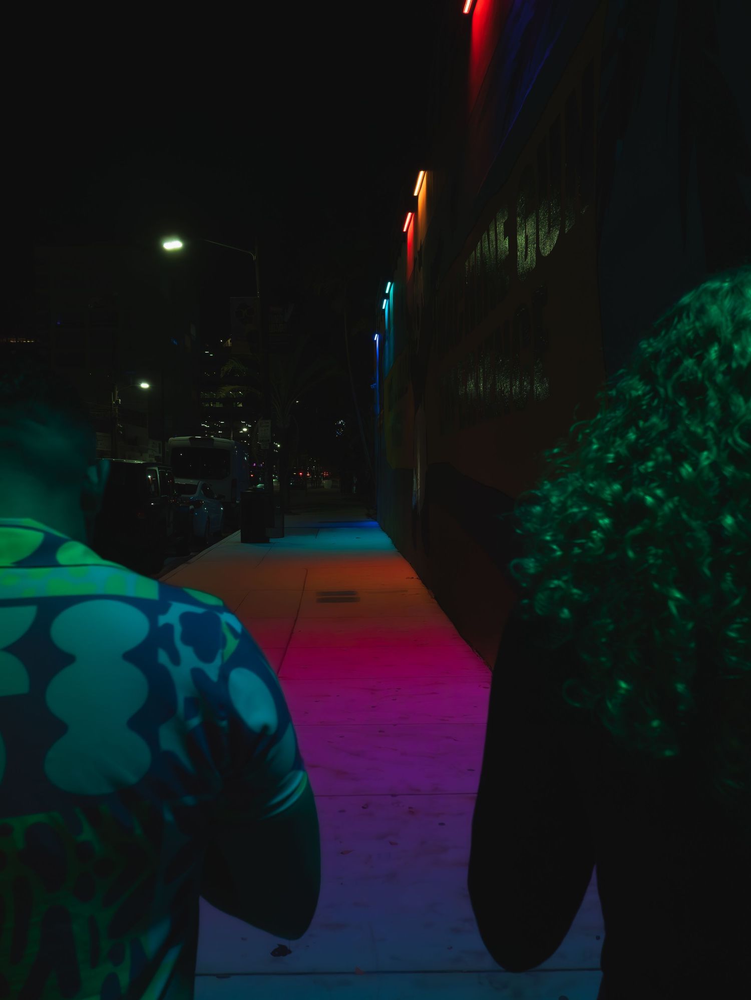 Colorful neon lights illuminate a dark street at night casting vibrant purple and green glows against the pavement.