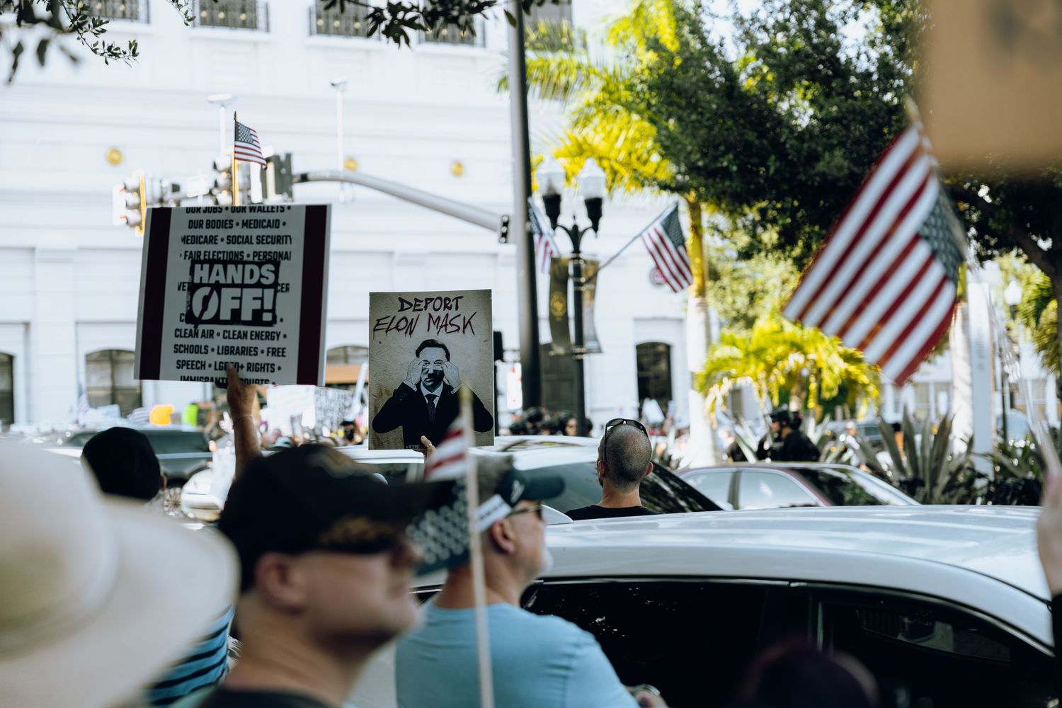 Protesters march down a city street while holding signs near parked cars and buildings.