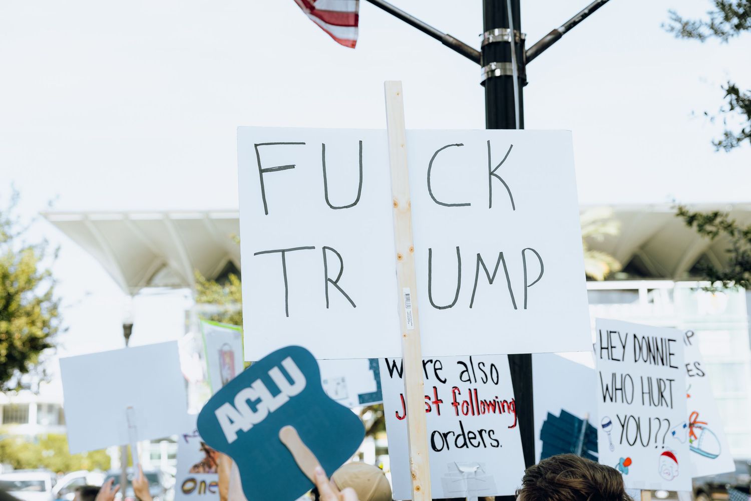 Protesters at political rally hold signs expressing opposition to Trump and carrying ACLU banners.