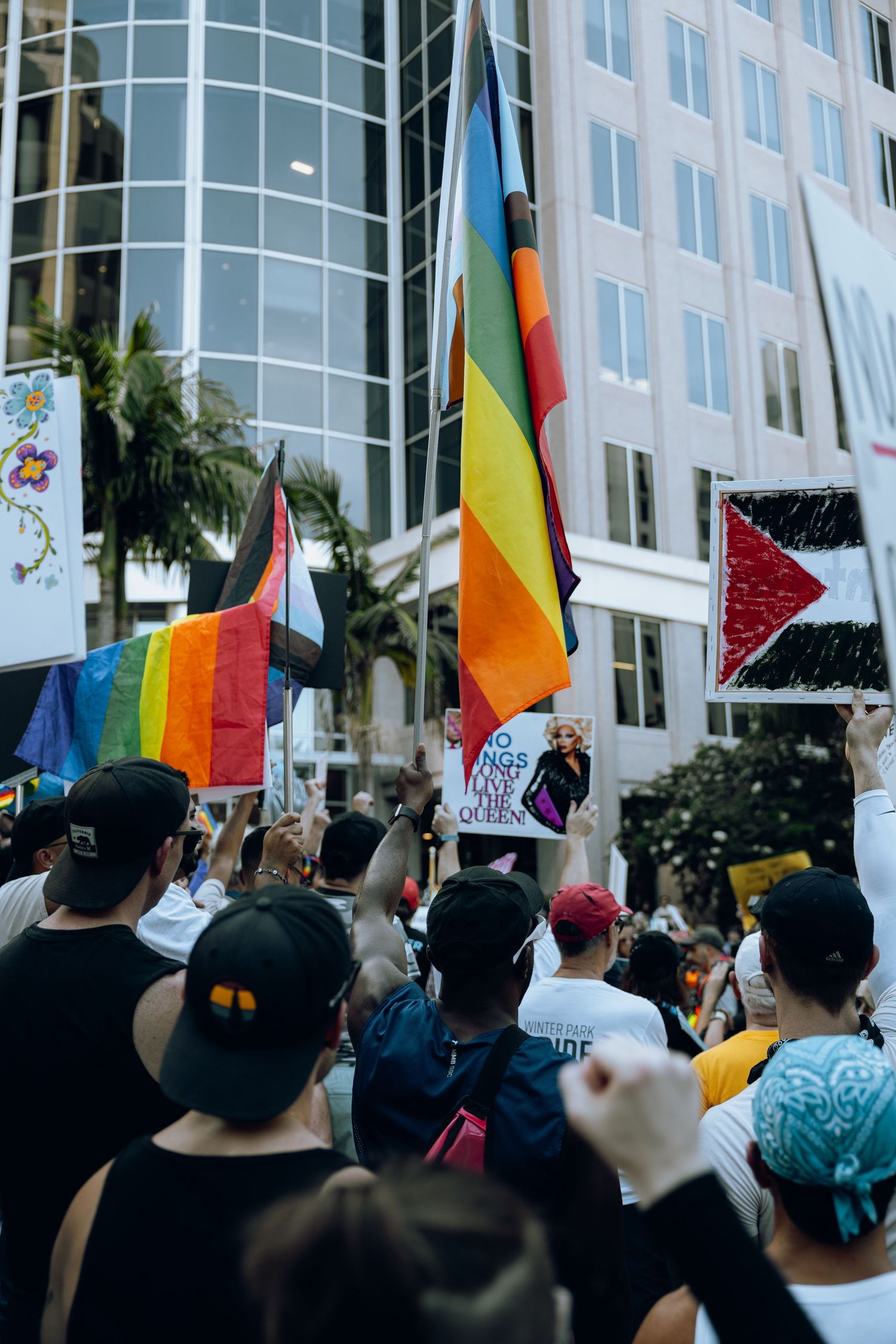 Rainbow flags and protest signs raised against modern urban building backdrop.