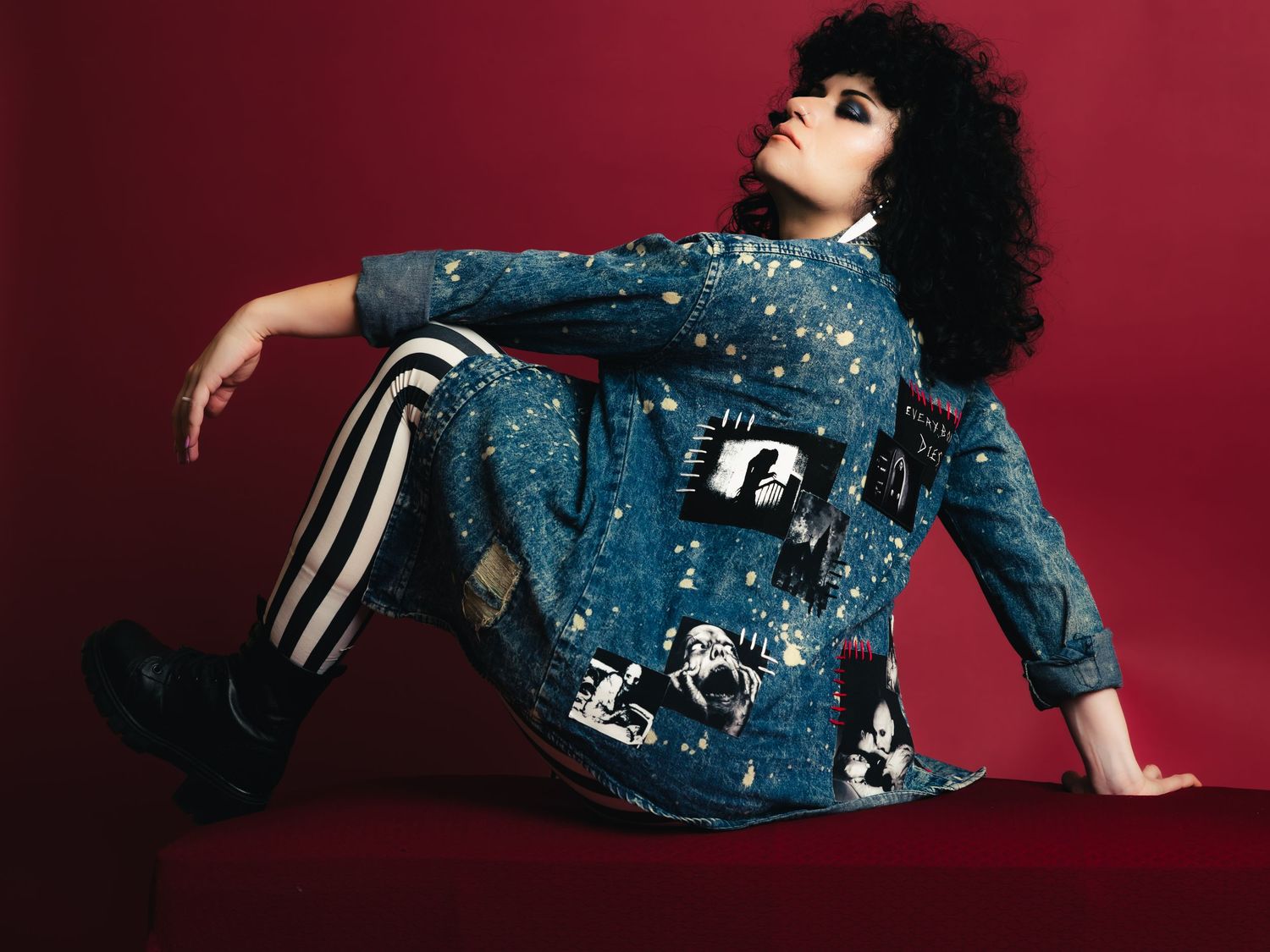 Model poses in decorated denim jacket, striped pants, and black boots against a red background.